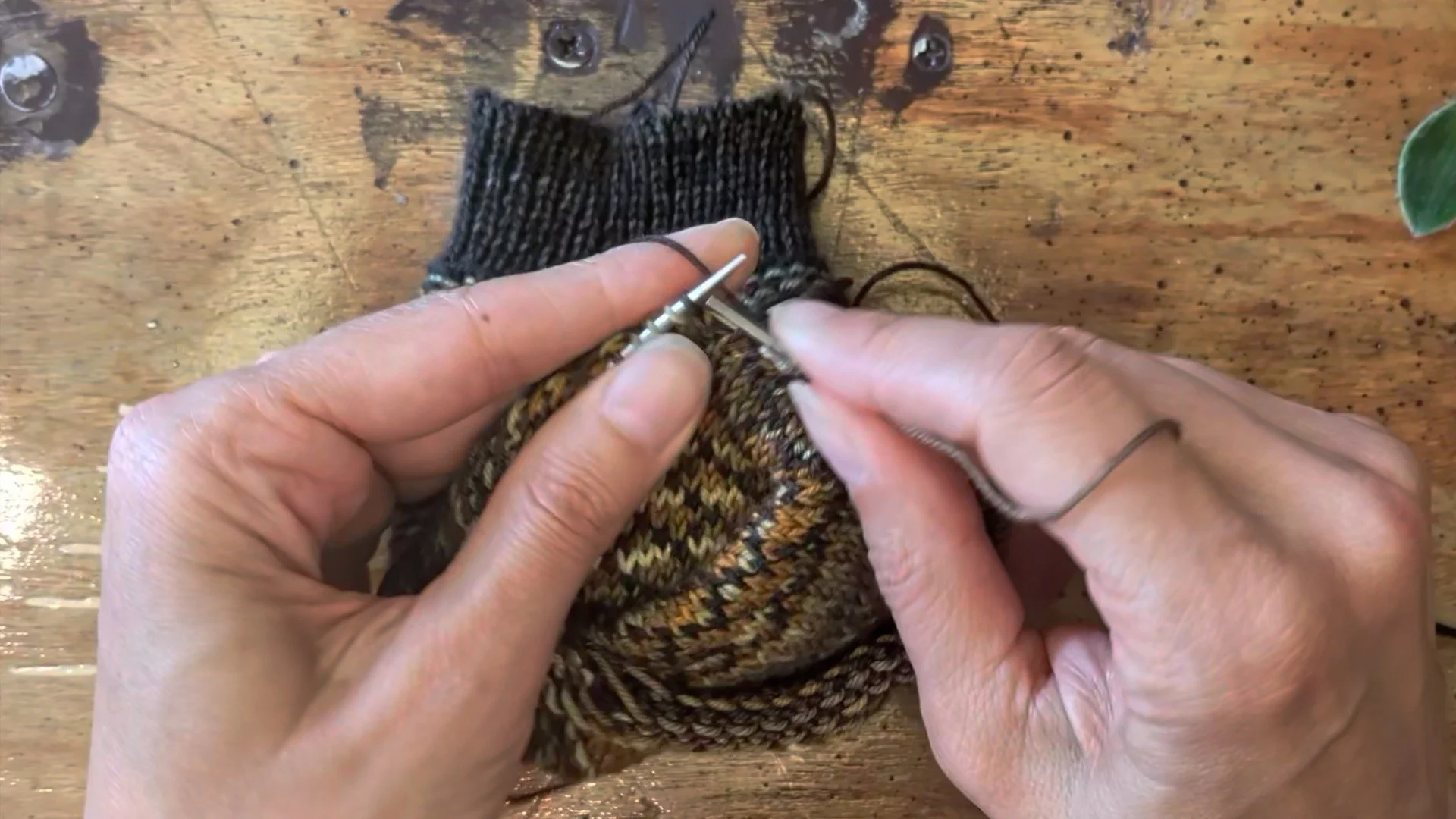 two hands are photographed kniitting a sock inside out on a wooden table.