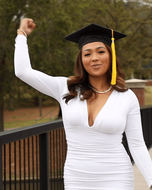 A young woman wearing a white fitted dress and a graduation cap with a yellow tassel, celebrating outdoors with her arm raised in a fist pump.