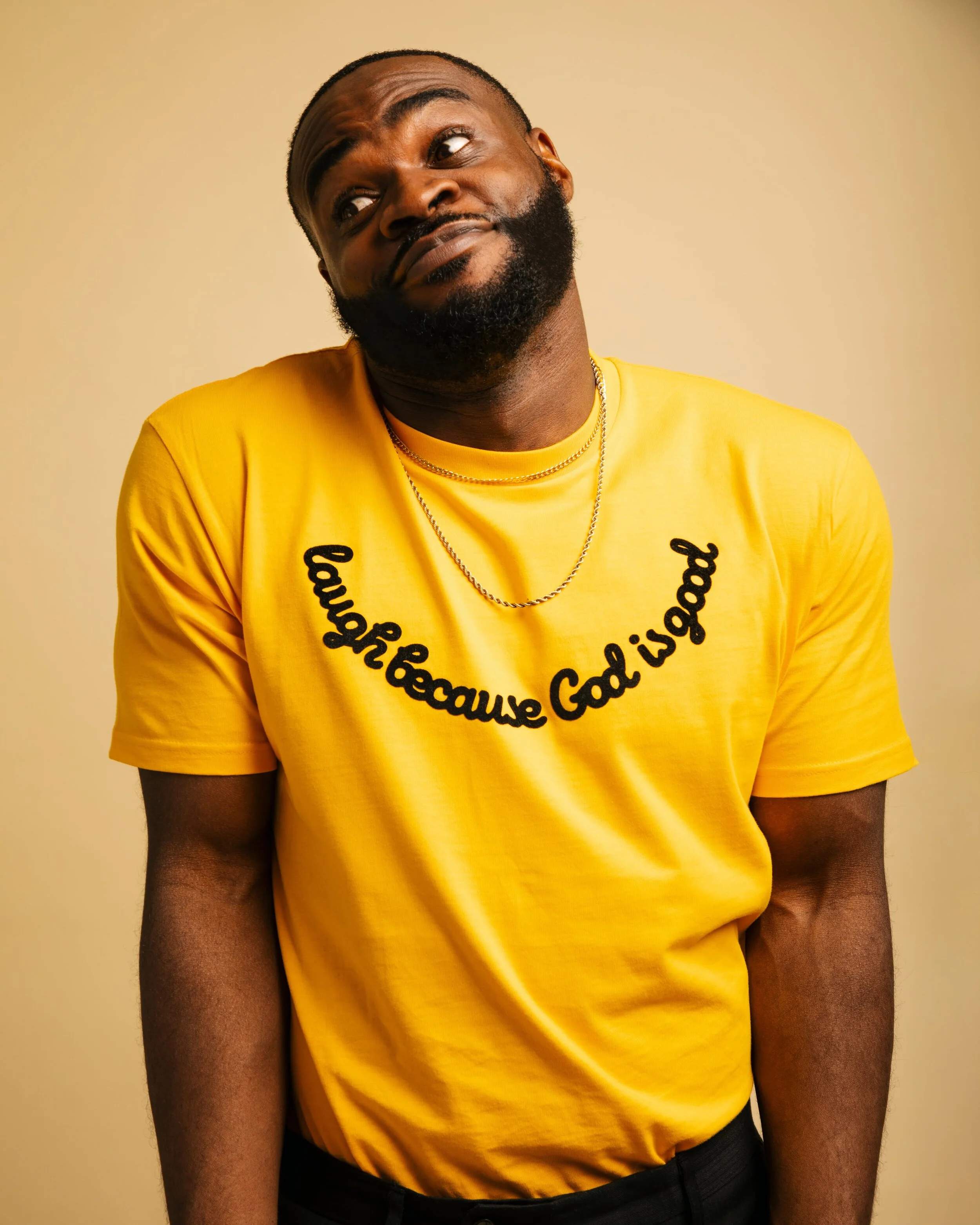 A man with a beard wearing a yellow t-shirt with black text and black pants, standing against a beige background.