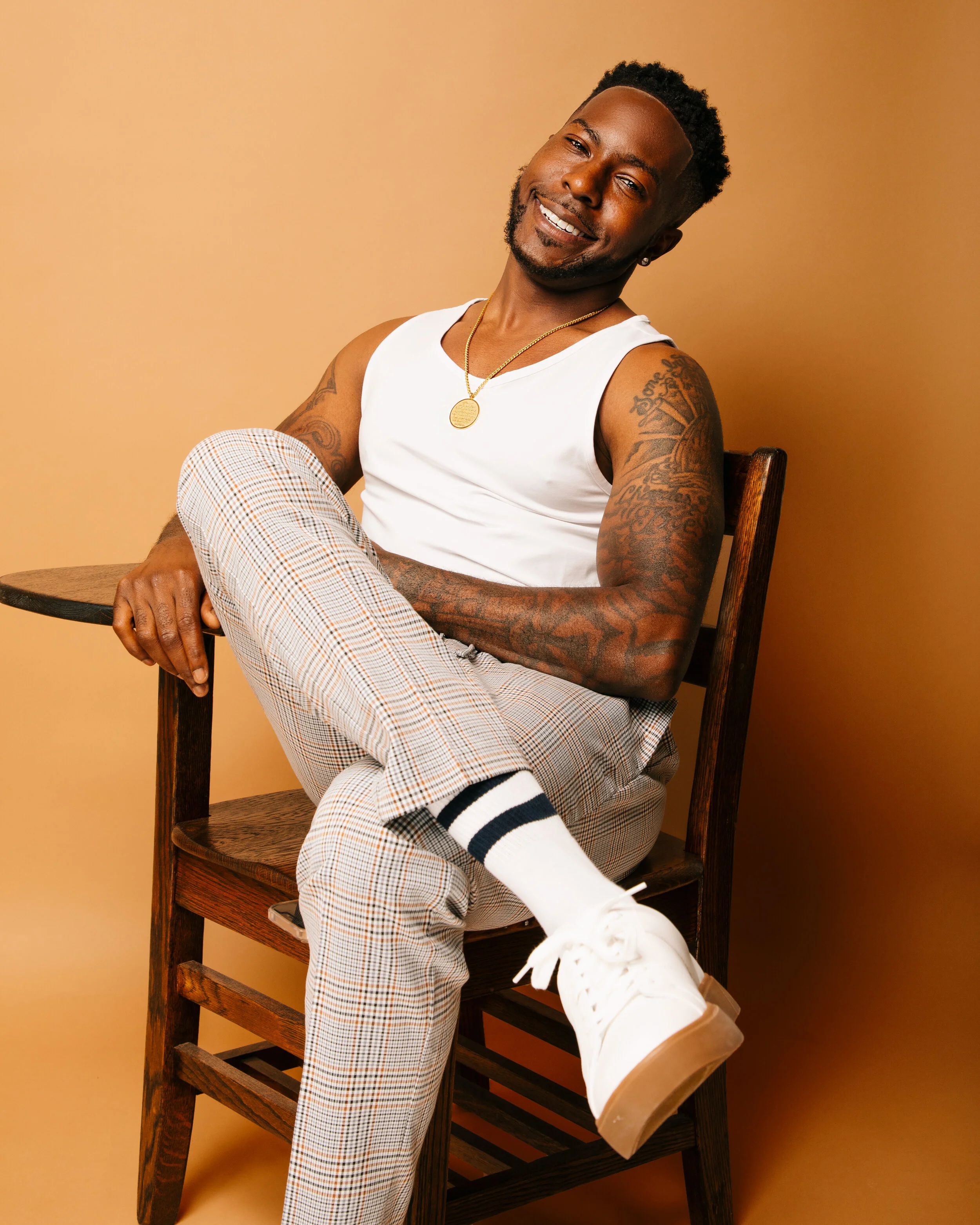 A smiling man with tattoos on his arms sitting on a wooden chair, wearing a white sleeveless top, plaid pants, white sneakers, a gold chain, and earrings, against a beige background.