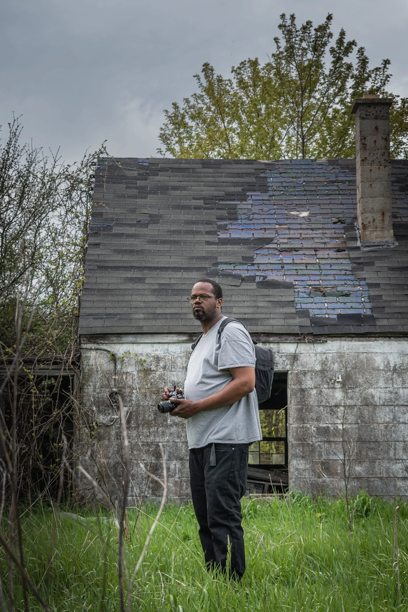 A man standing in a grassy area holding a camera, in front of a dilapidated house with a damaged roof and a rusty chimney, surrounded by overgrown vegetation and trees under an overcast sky.