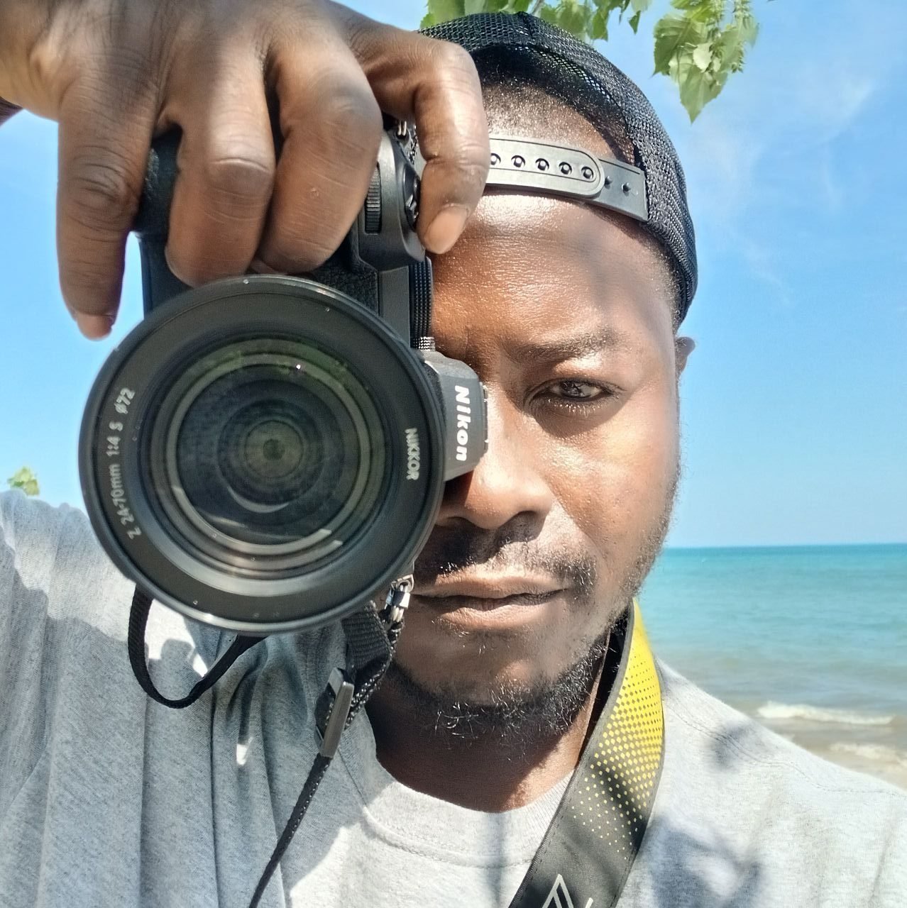 A man holding a Nikon camera takes a selfie at the beach with blue sky, green leaves, and ocean in the background.