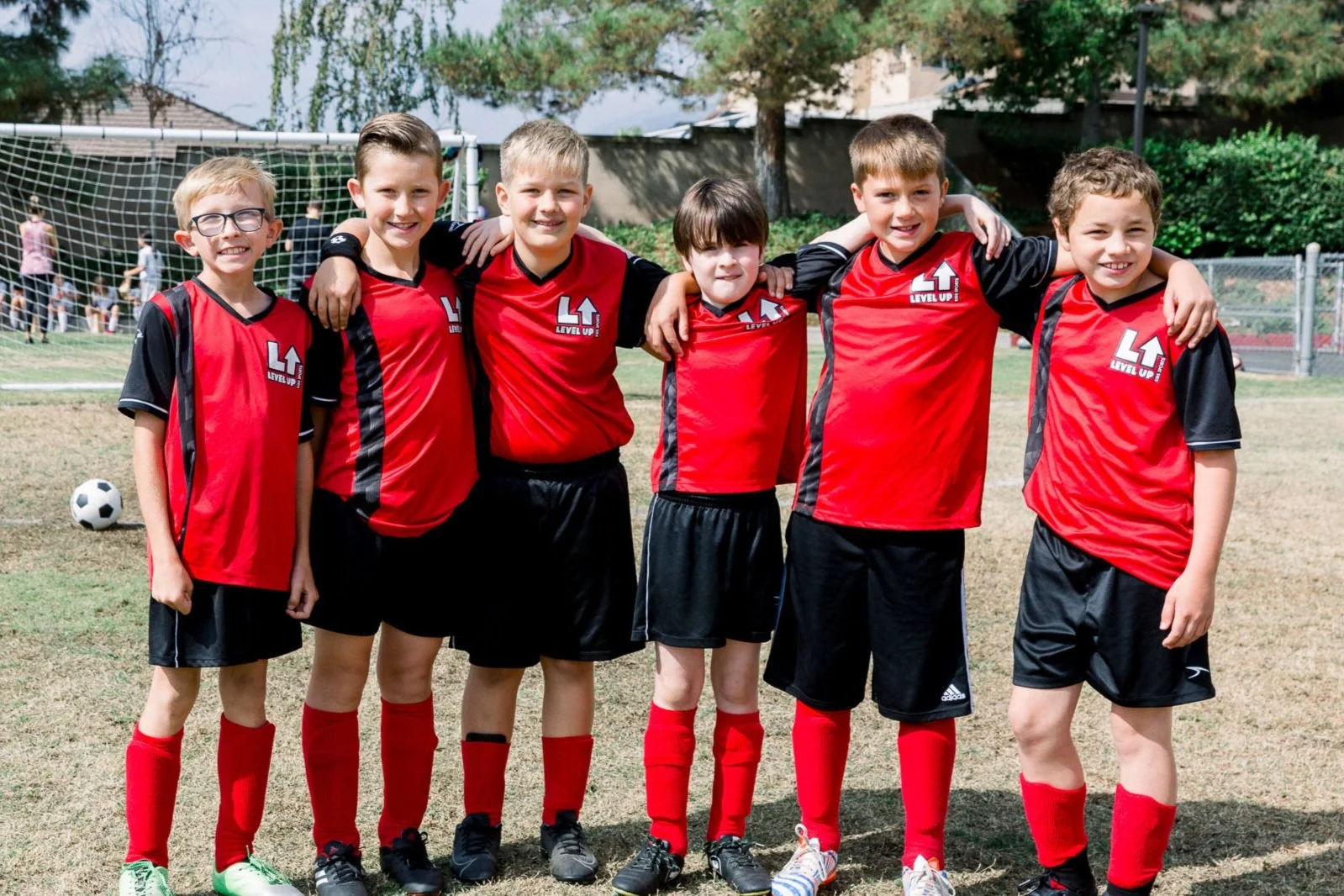 Group of seven young boys in red and black soccer uniforms standing together on a soccer field, front of a goalpost, with trees and other children in the background.