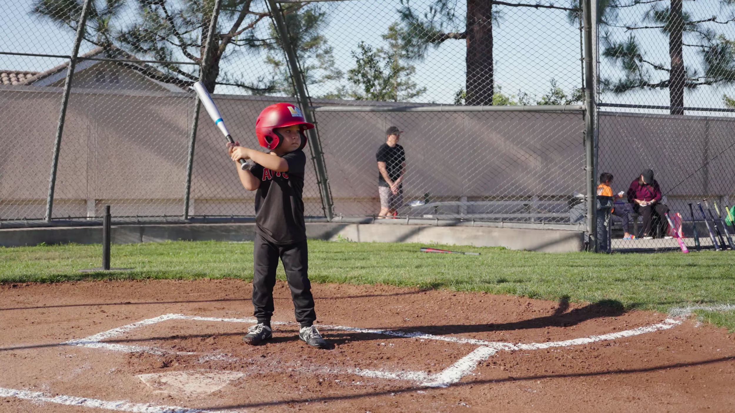 Young boy in a red baseball jersey and gray pants practicing batting in a baseball or softball game, using a tee to hit the ball. He is wearing a helmet and appears focused. In the background, spectators are seated and standing behind a chain-link fence, with baseball equipment visible.
