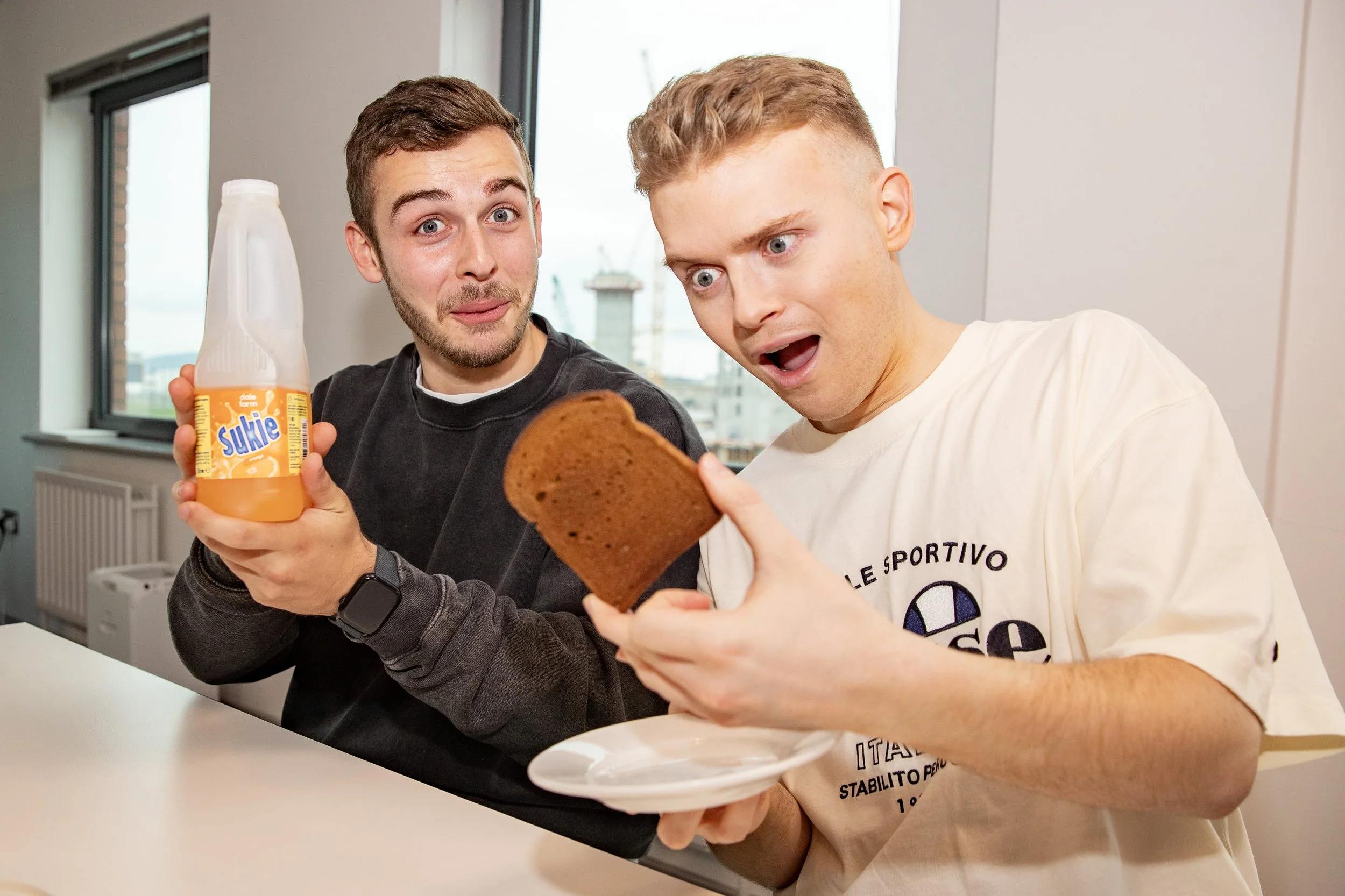Two young men sitting at a table with surprised expressions, one holding a slice of bread and the other holding a bottle of orange juice.