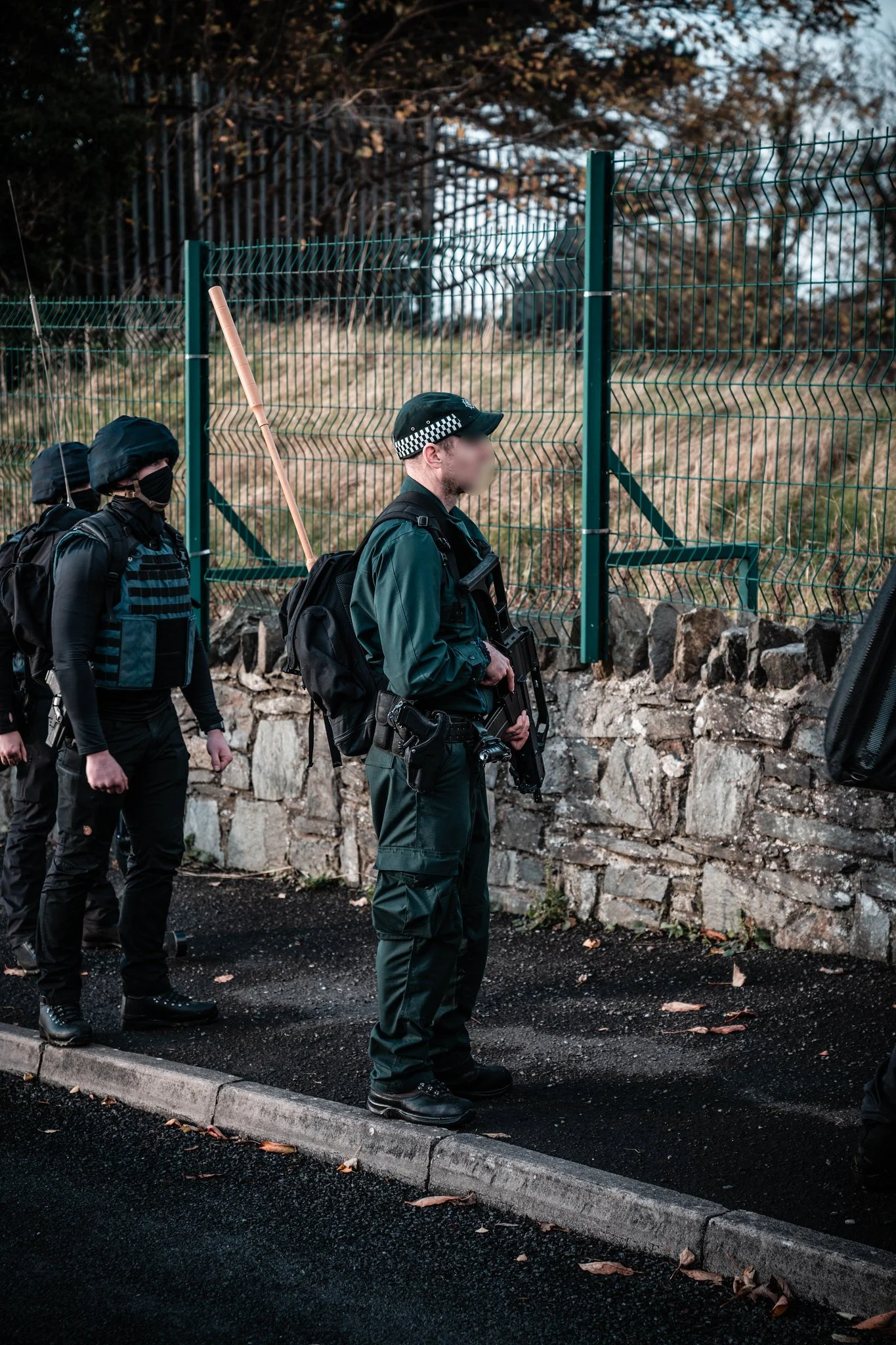 Two police officers in tactical gear standing on a sidewalk near a green metal fence, with one officer holding a firearm, during daytime.