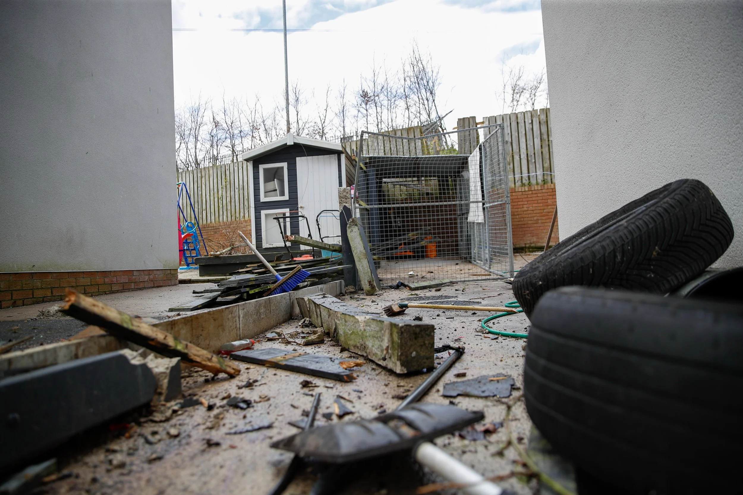 Debris and damaged items scattered on the ground outside, with a small shed and a fence in the background, and a pile of tires on the right side.