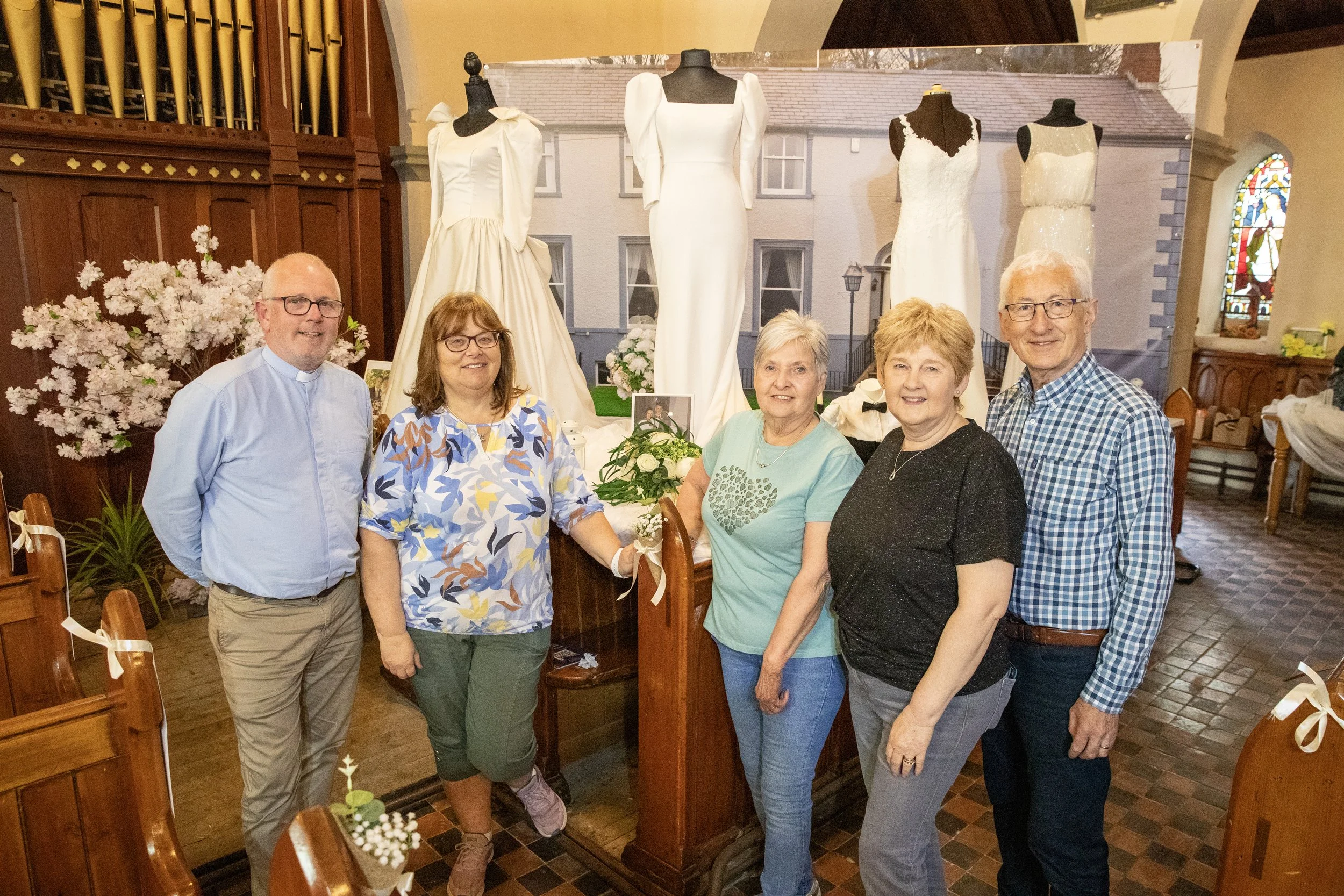 Group of five people standing inside a church, celebrating a wedding with four wedding dresses displayed on mannequins behind them.