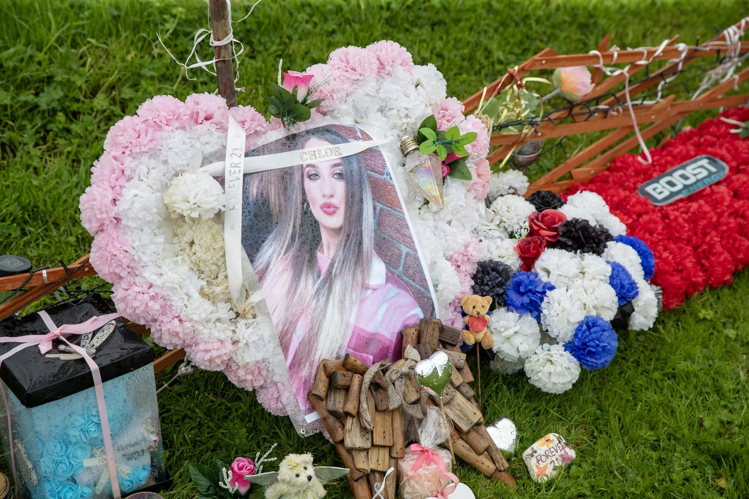 A funeral memorial on the grass with a large pink and white flower wreath, a picture of a young woman with long hair, and various small stuffed animals and decorative items.