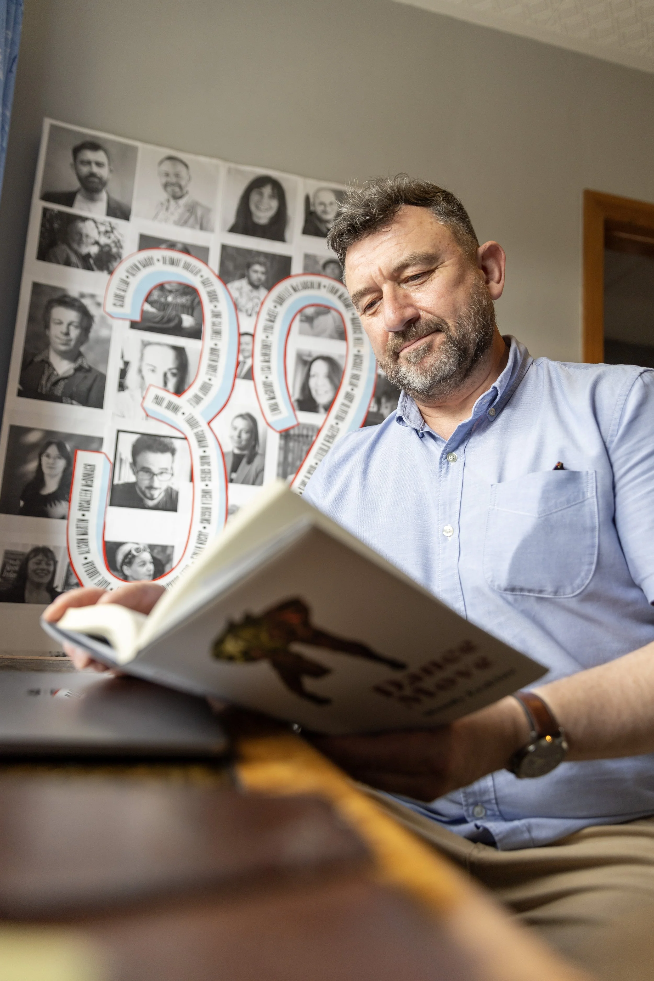 A bearded man in a light blue shirt reads a book at a desk; a collage of black and white photos with a large red and white 