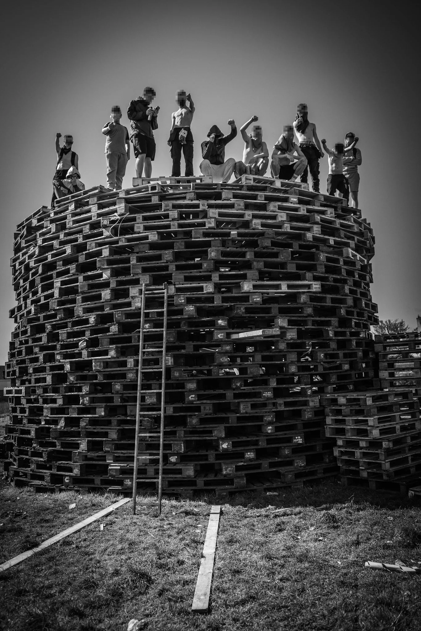 Group of people standing atop a tall stack of wooden pallets outdoors, some shirtless, raising their fists or holding objects, in black and white.