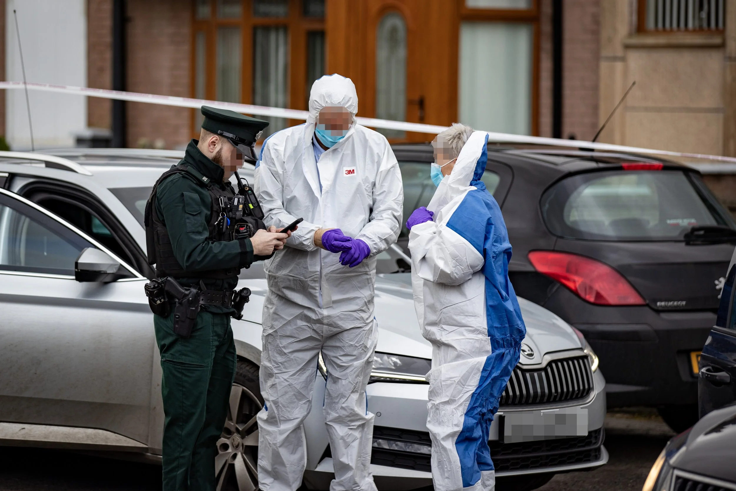 Police officers and forensic investigators at a crime scene on a city street, standing next to parked cars, with one officer looking at a mobile device and the investigators wearing protective suits and masks.