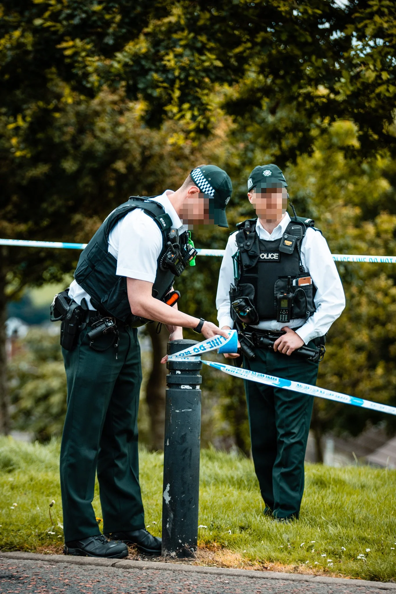Two police officers in uniform and tactical vests are working at a crime scene, sealing off the area with police tape outdoors near trees and grass.