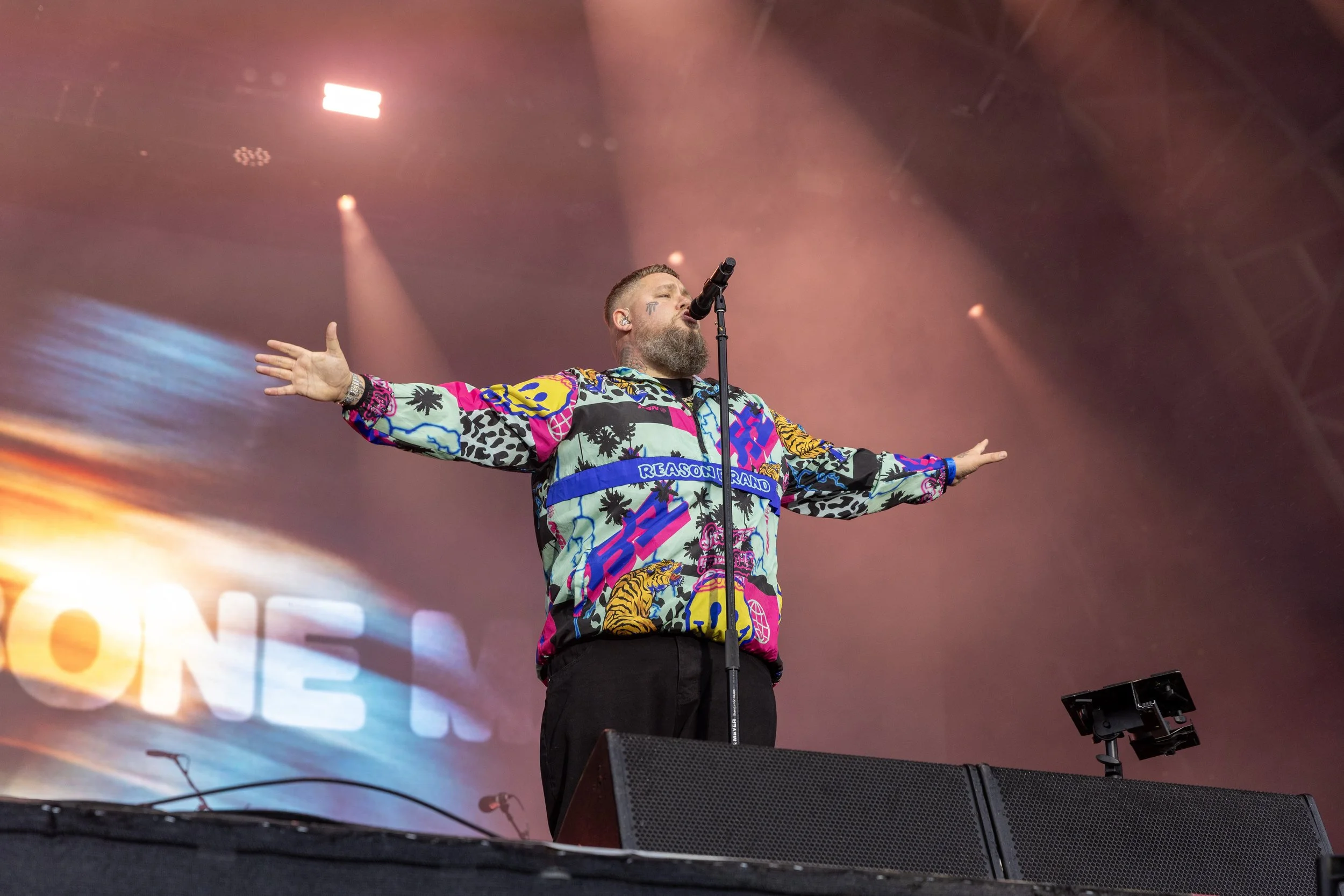 A man with a beard and tattoos singing into a microphone on stage, wearing a colorful, patterned jacket, with arms outstretched and stage lights shining behind him.