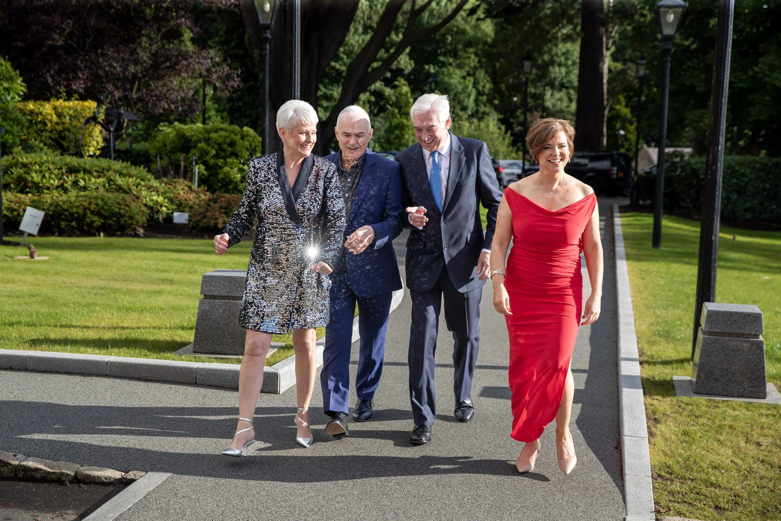 Four well-dressed adults, three men and one woman, walking along a park pathway, smiling and laughing on a sunny day.