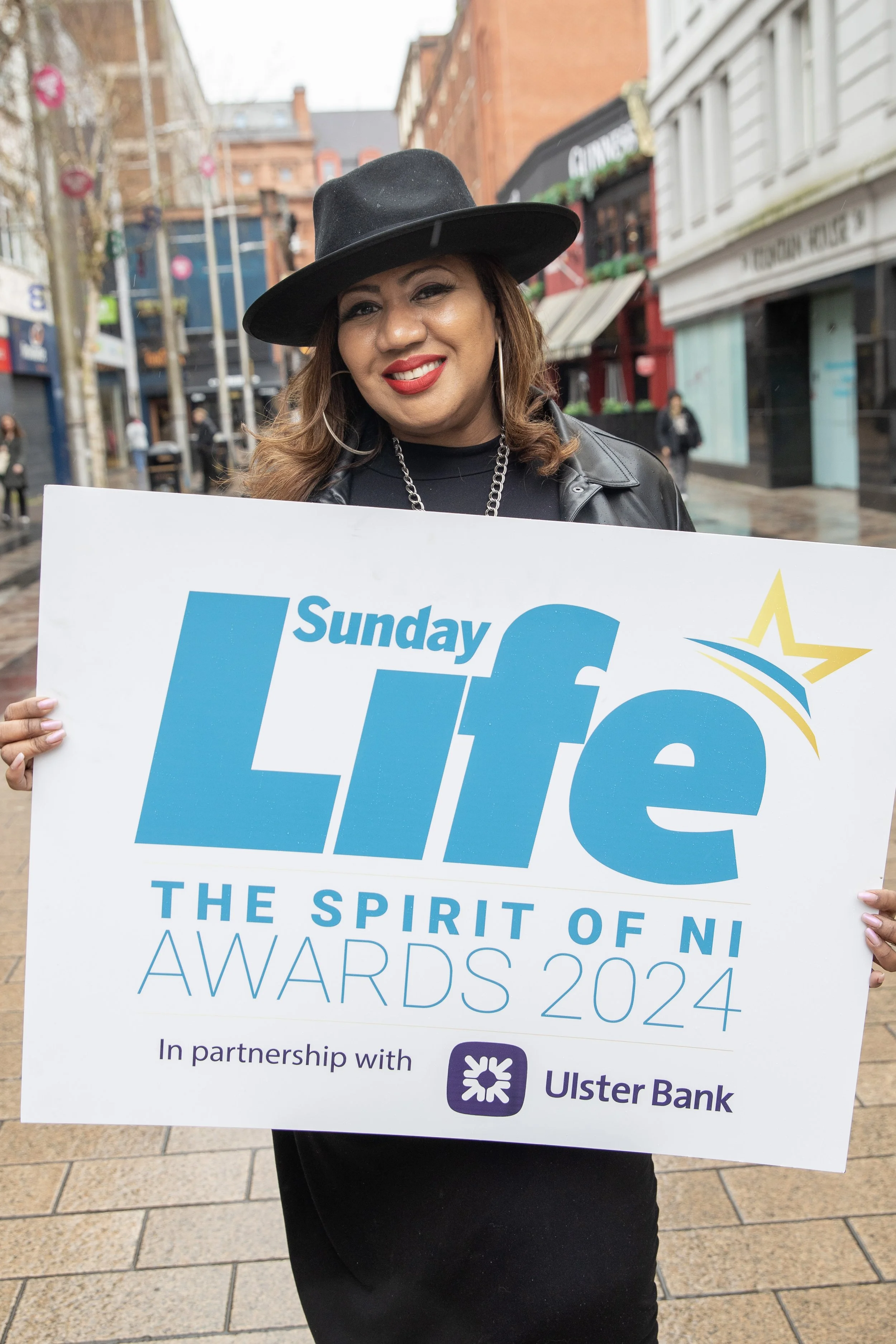 A woman in a black hat and leather jacket smiling and holding a sign for the Sunday Life Awards 2024, in partnership with Ulster Bank, outdoors in an urban area.