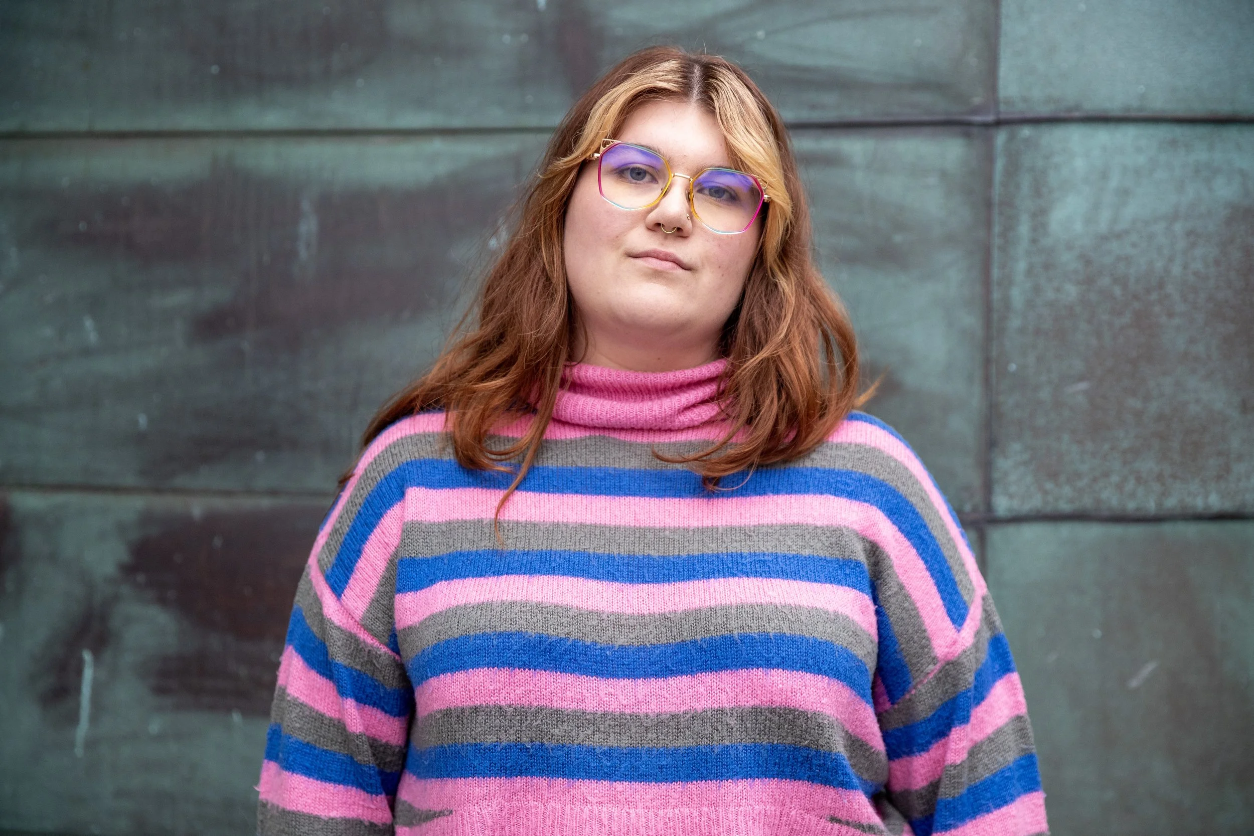 A woman with red hair wearing colorful striped sweater and pink glasses standing in front of a dark green wall.