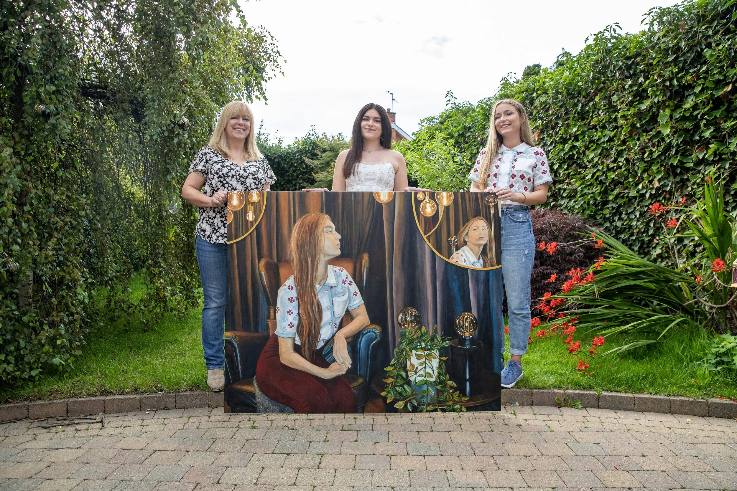 Three women standing outdoors in a garden holding a large painting that depicts a woman sitting on a chair with long hair, wearing a flower-patterned shirt, and seen in two different angles with dark curtains and string lights in the background.
