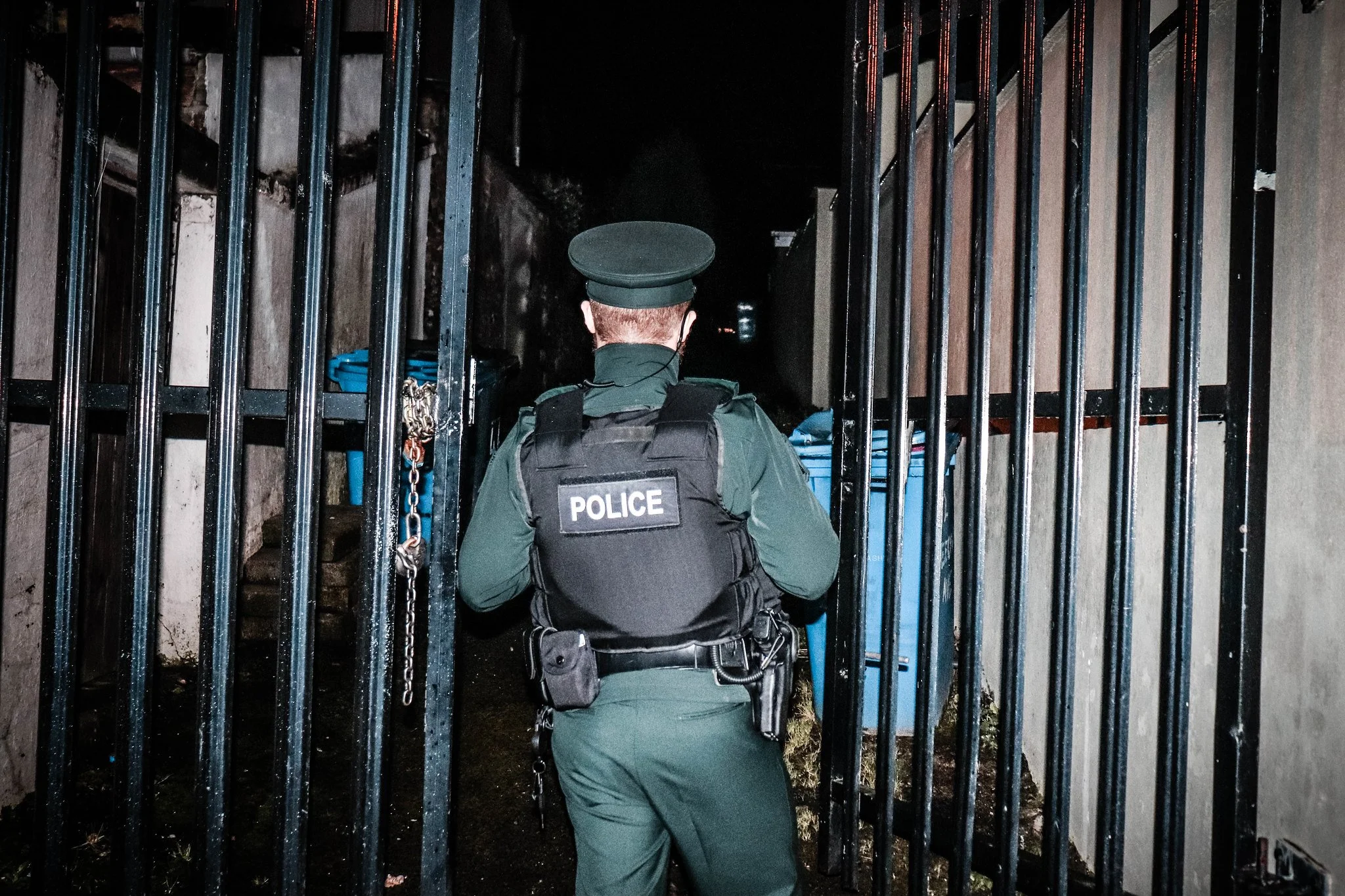 Police officer in uniform and vest walking through black metal gate at night.