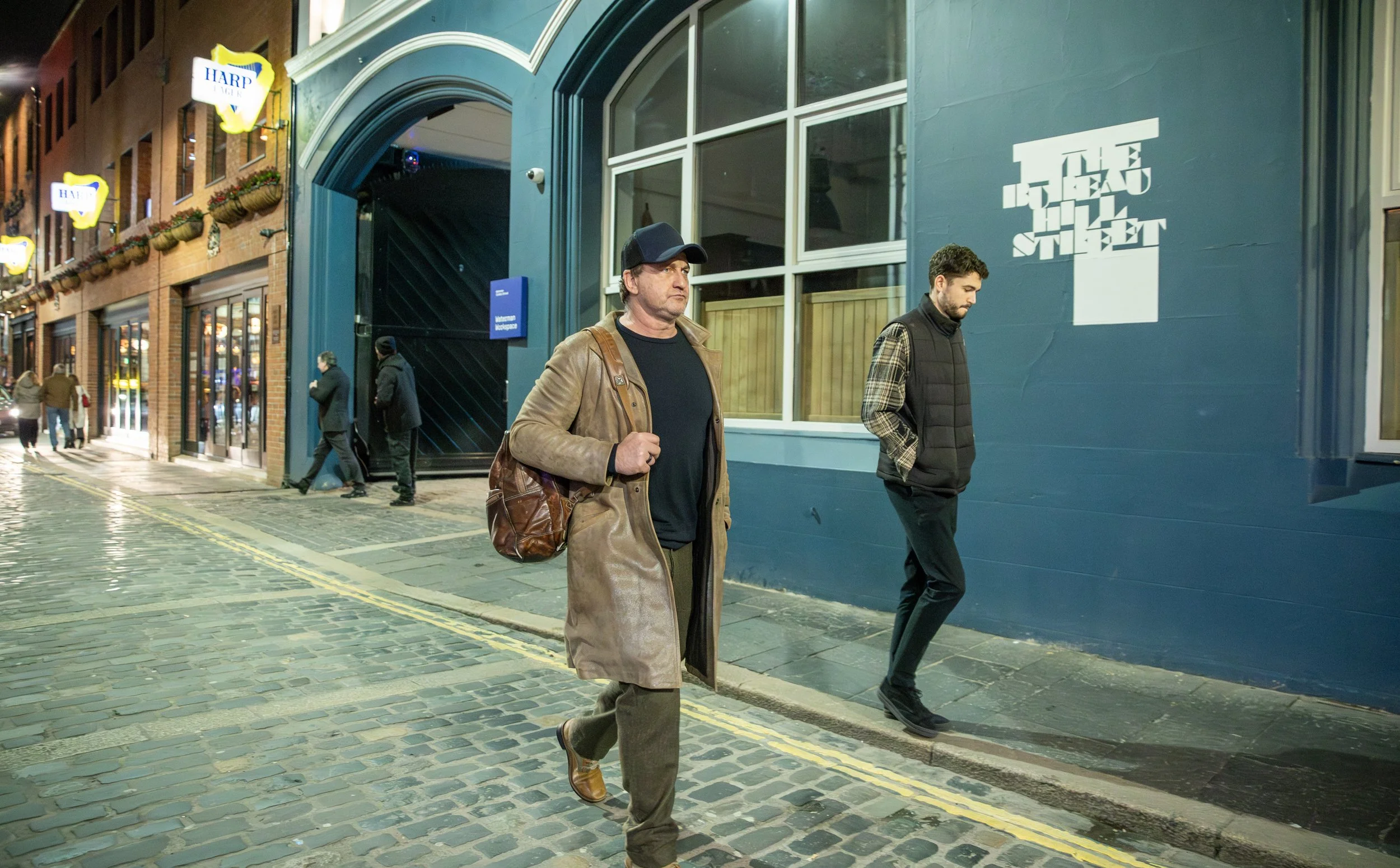 Two men walking on a cobblestone sidewalk at night in front of a blue building with a large computer monitor logo on the wall. One man wears a brown trench coat and baseball cap, the other wears a black padded vest over a plaid shirt.