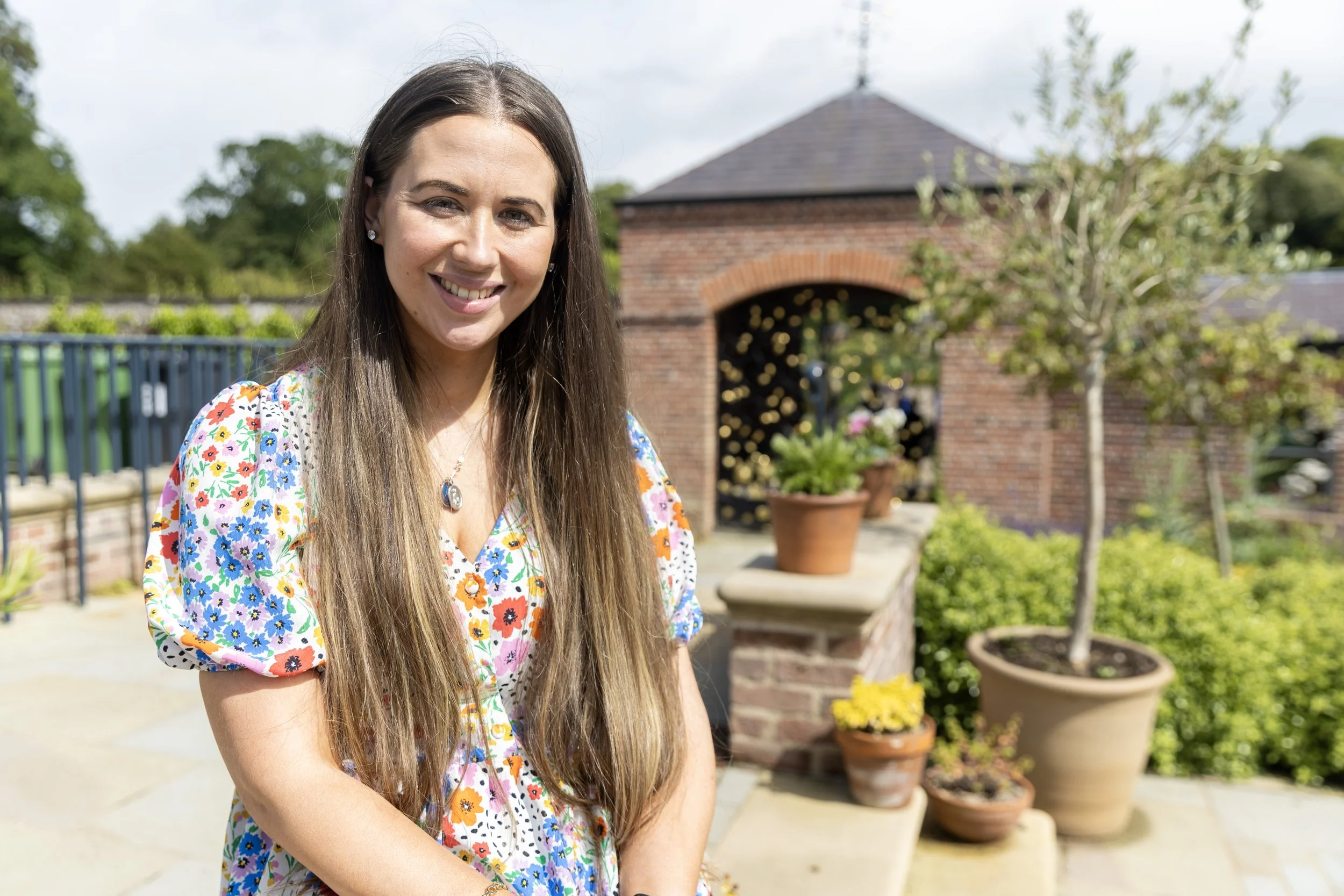 A woman with long brown hair smiling outdoors in front of a garden with potted plants, brick structures, and a tree, on a partly cloudy day.