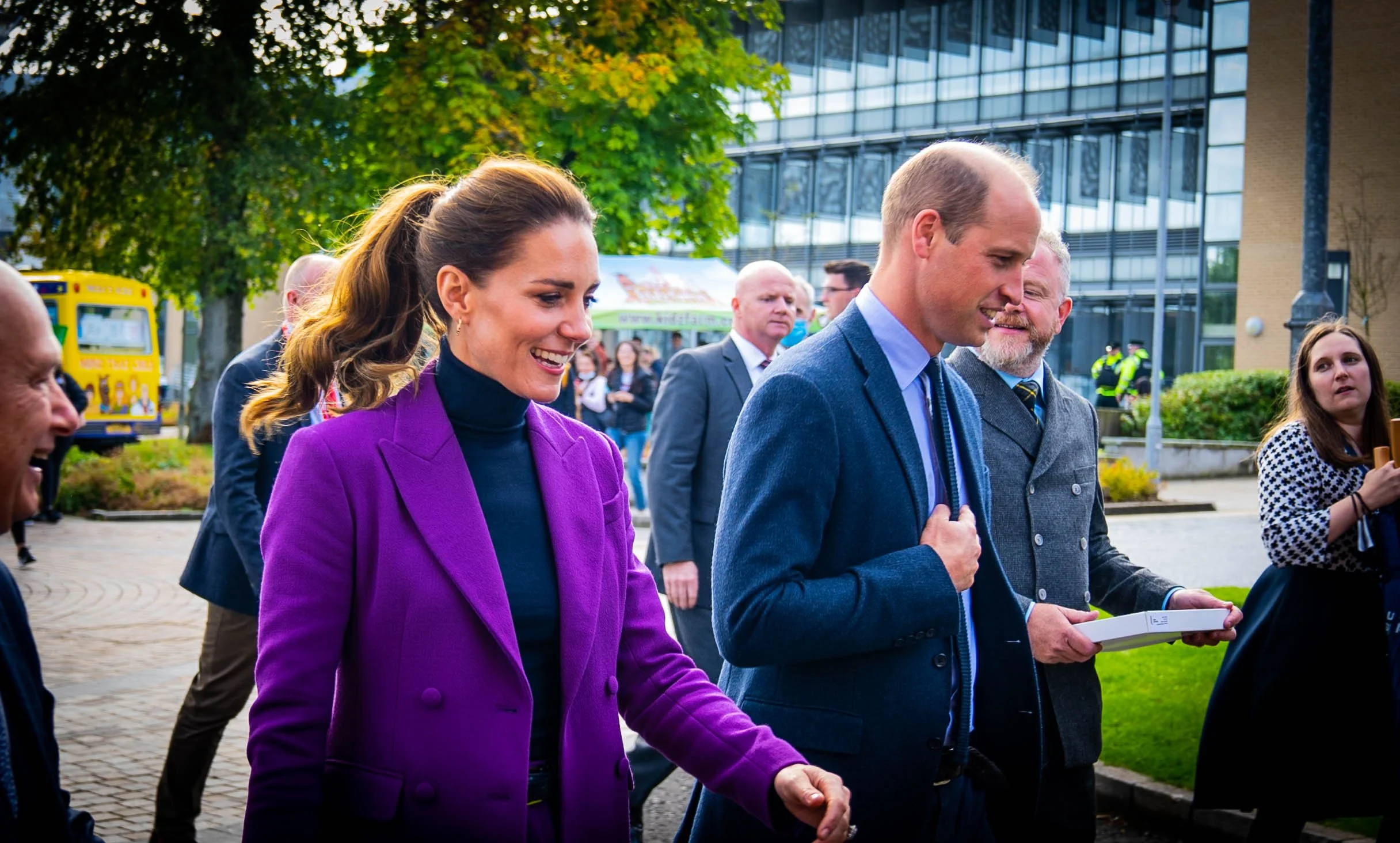 A group of well-dressed people, including a woman in a purple blazer and a man in a suit, walking outdoors in front of a modern glass building with other people and a yellow bus in the background.