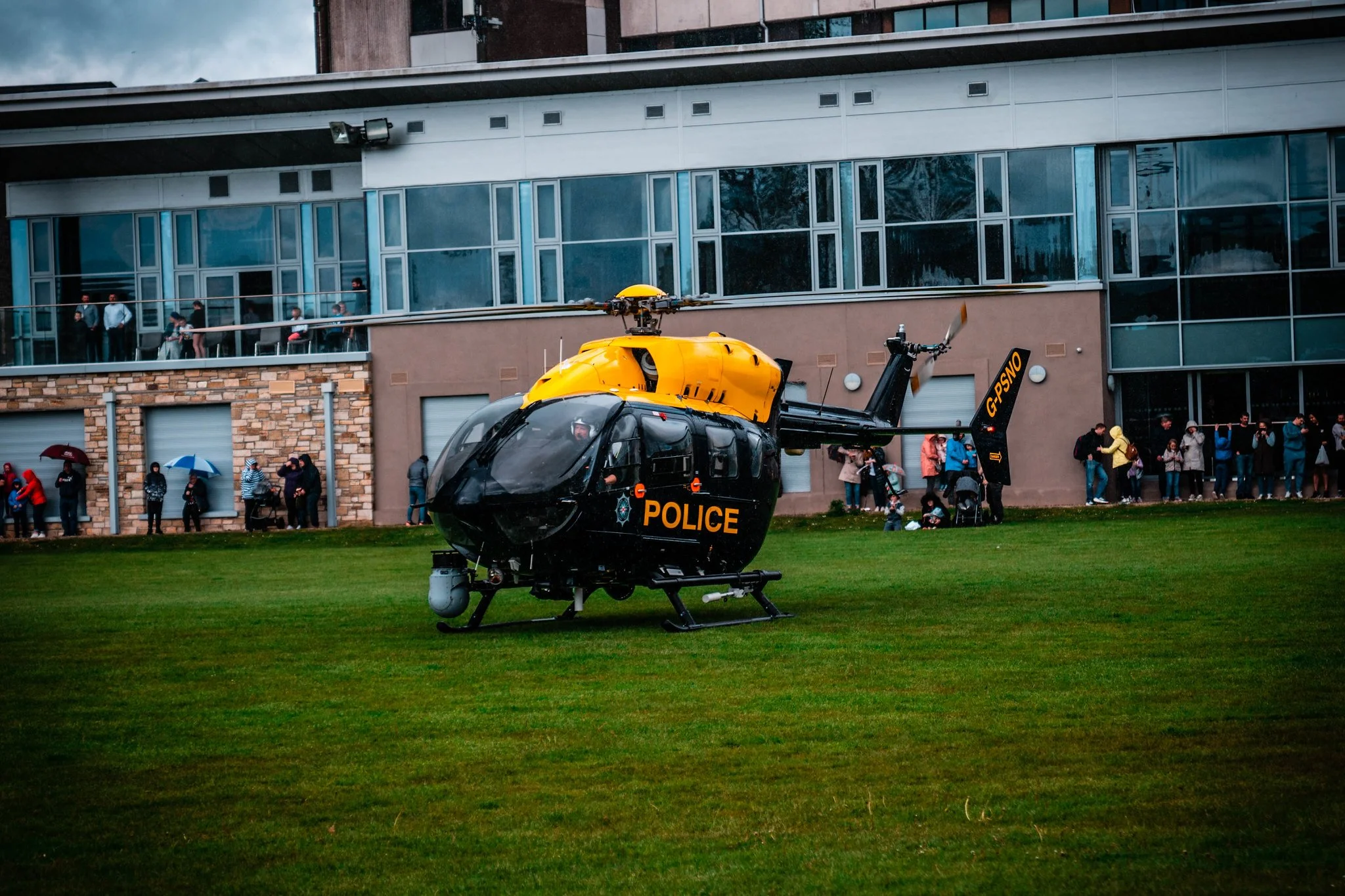 A black and yellow police helicopter on a grassy field with people standing and watching behind a building with large windows.