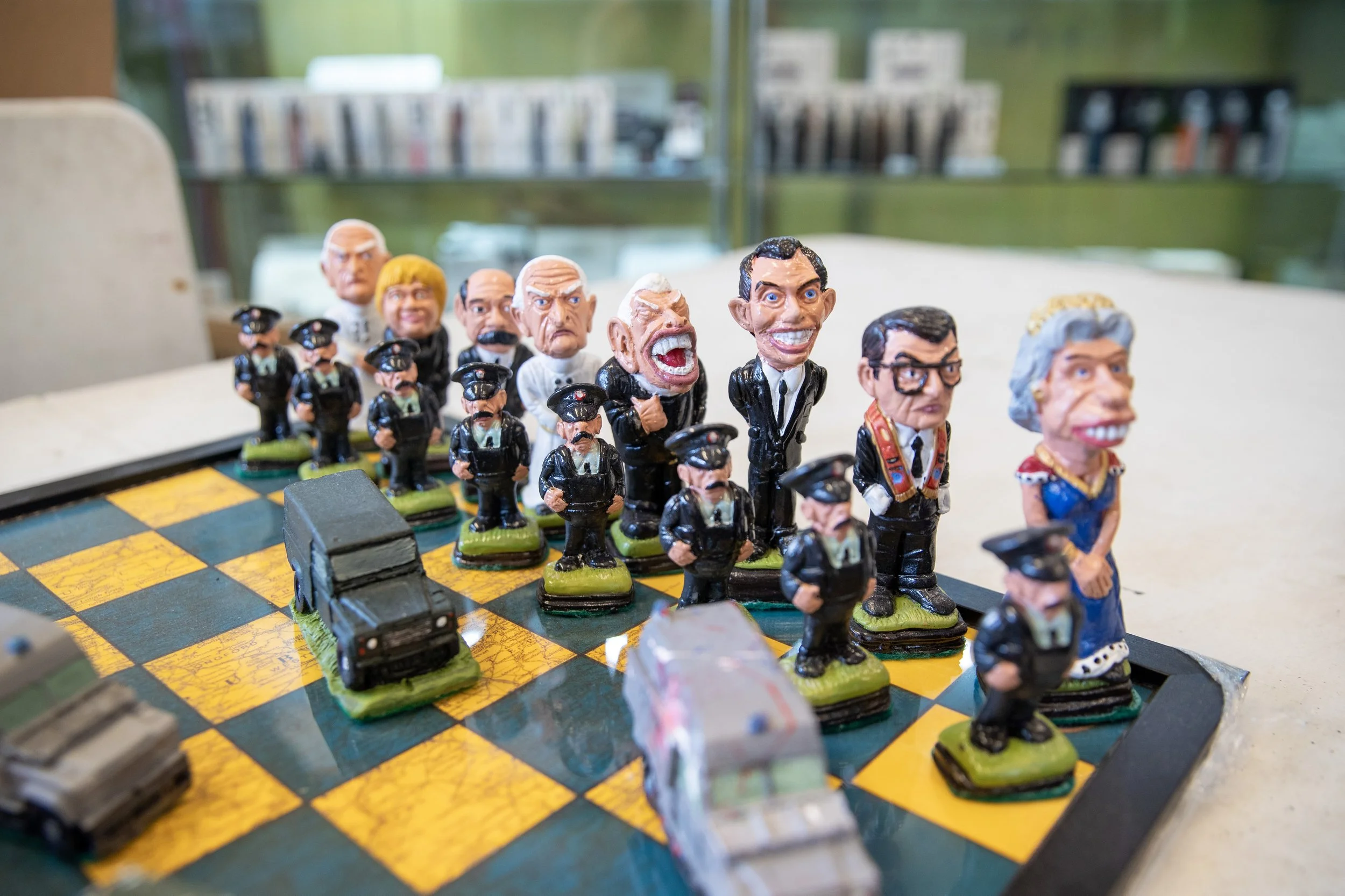 A chess set with pieces modeled as caricatured figures, including politicians and a queen, arranged on a yellow and black checkered board, with cars as part of the scene.