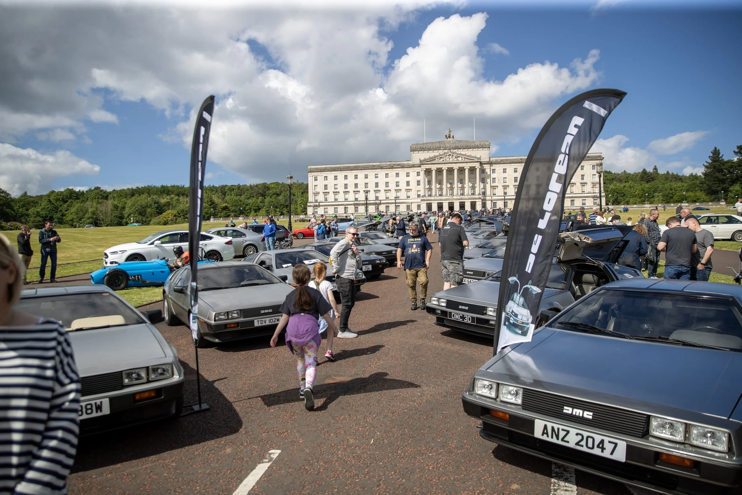 People attending a classic DeLorean car show in front of a historic building, with several DeLorean cars on display, flags, and a partly cloudy sky.