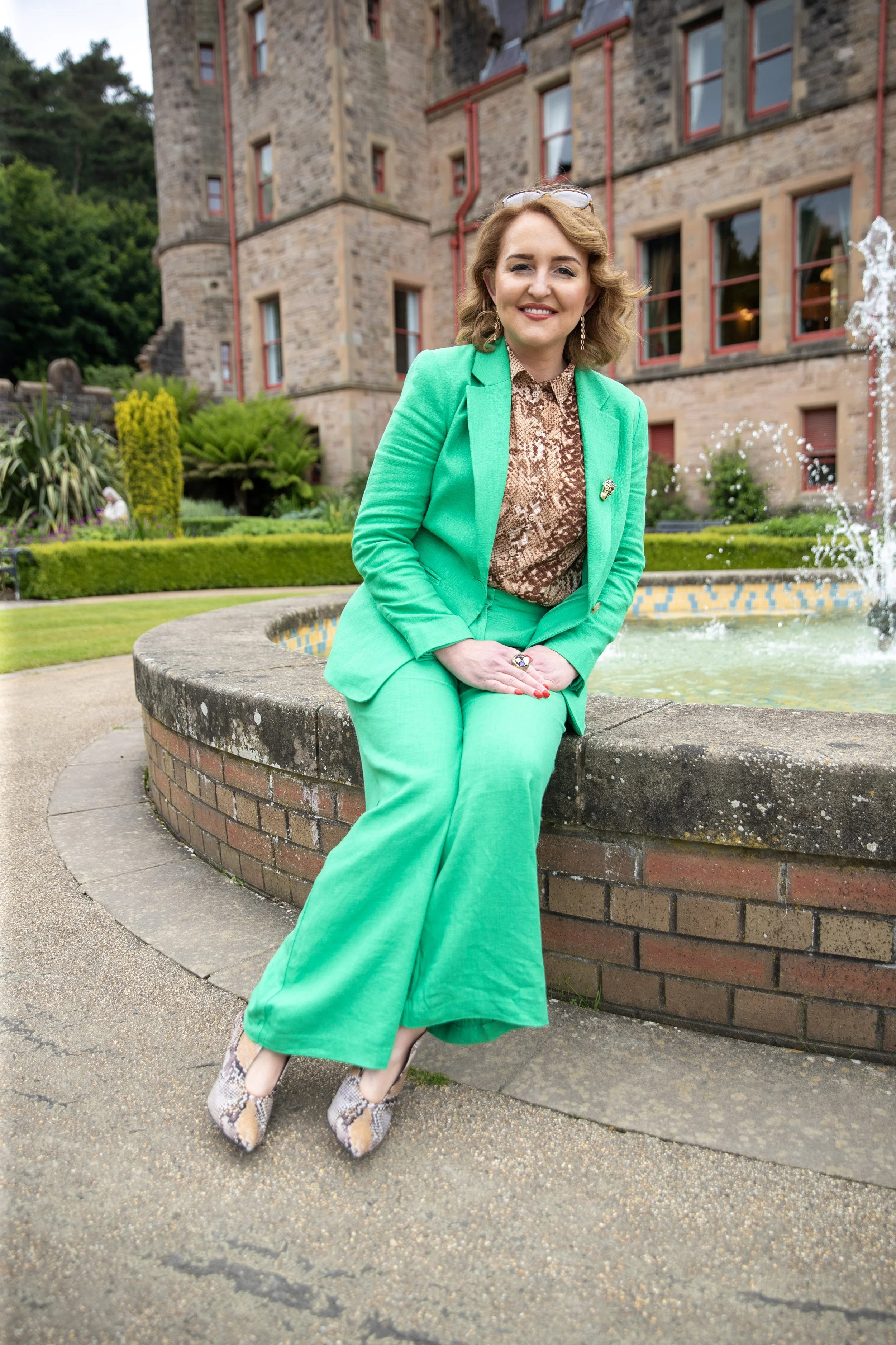 A woman wearing a bright green suit sitting on a brick curb in front of a fountain near a historic stone building with red-trimmed windows.