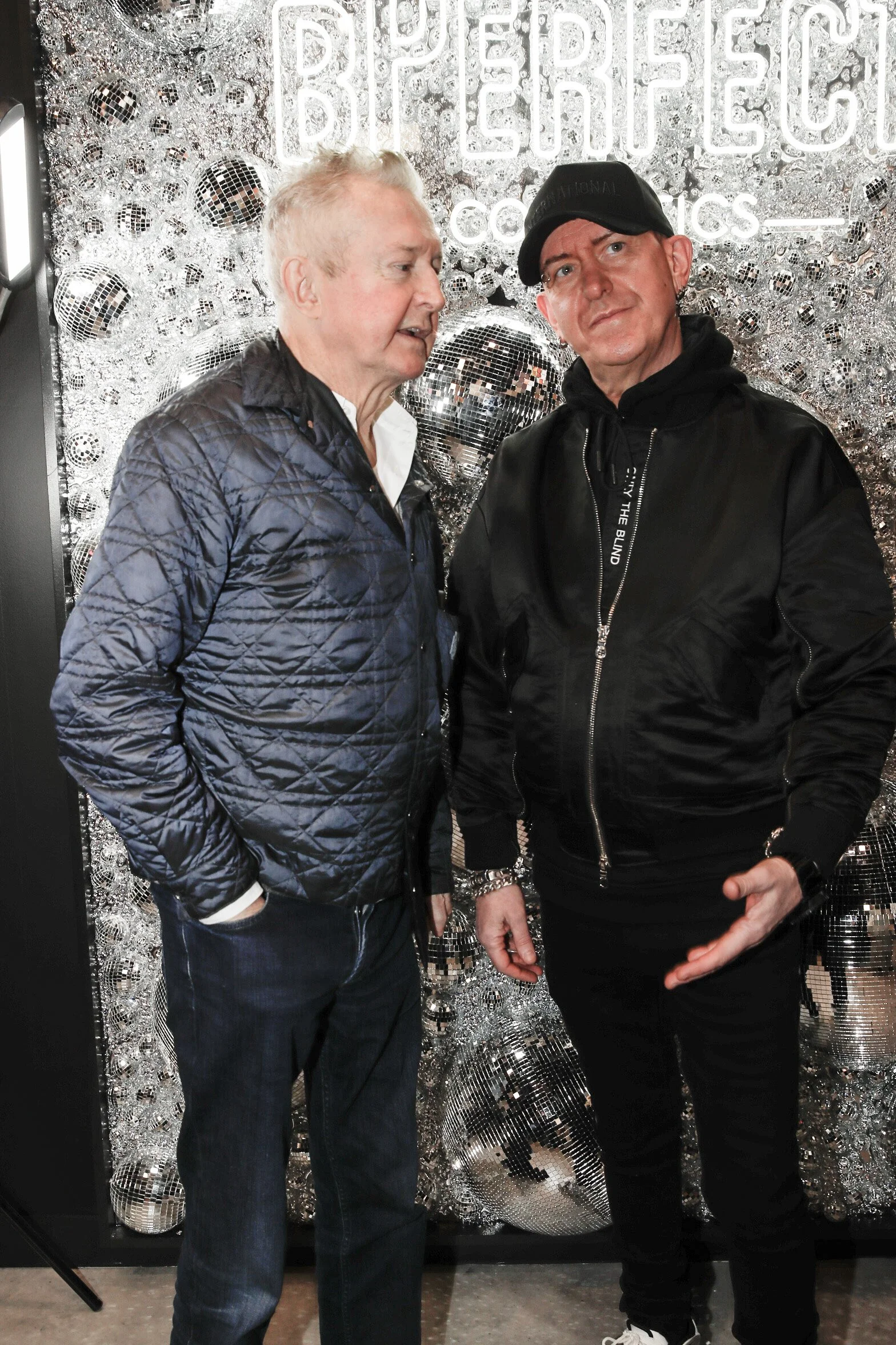 Two men standing in front of a shiny disco ball display with the text 'BE PERFECT' illuminated behind them. One man has white hair and is wearing a quilted navy jacket, the other is wearing a black cap and black jacket.