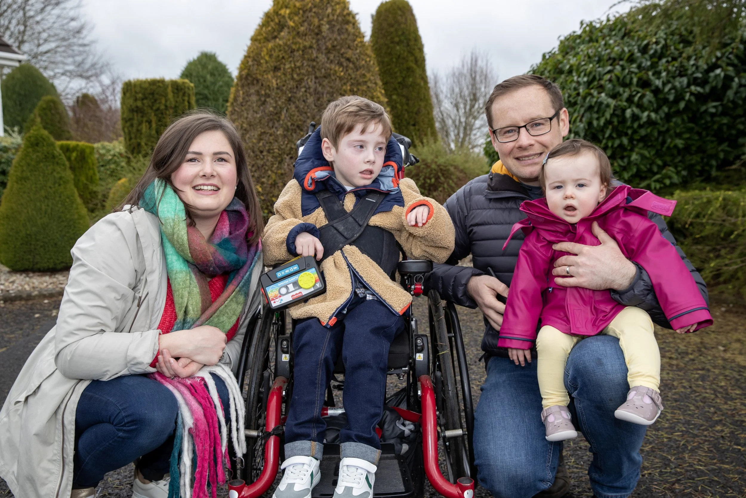 Family outdoors in a garden, smiling, with two children, one in a wheelchair, and a man holding a young girl, during daytime.