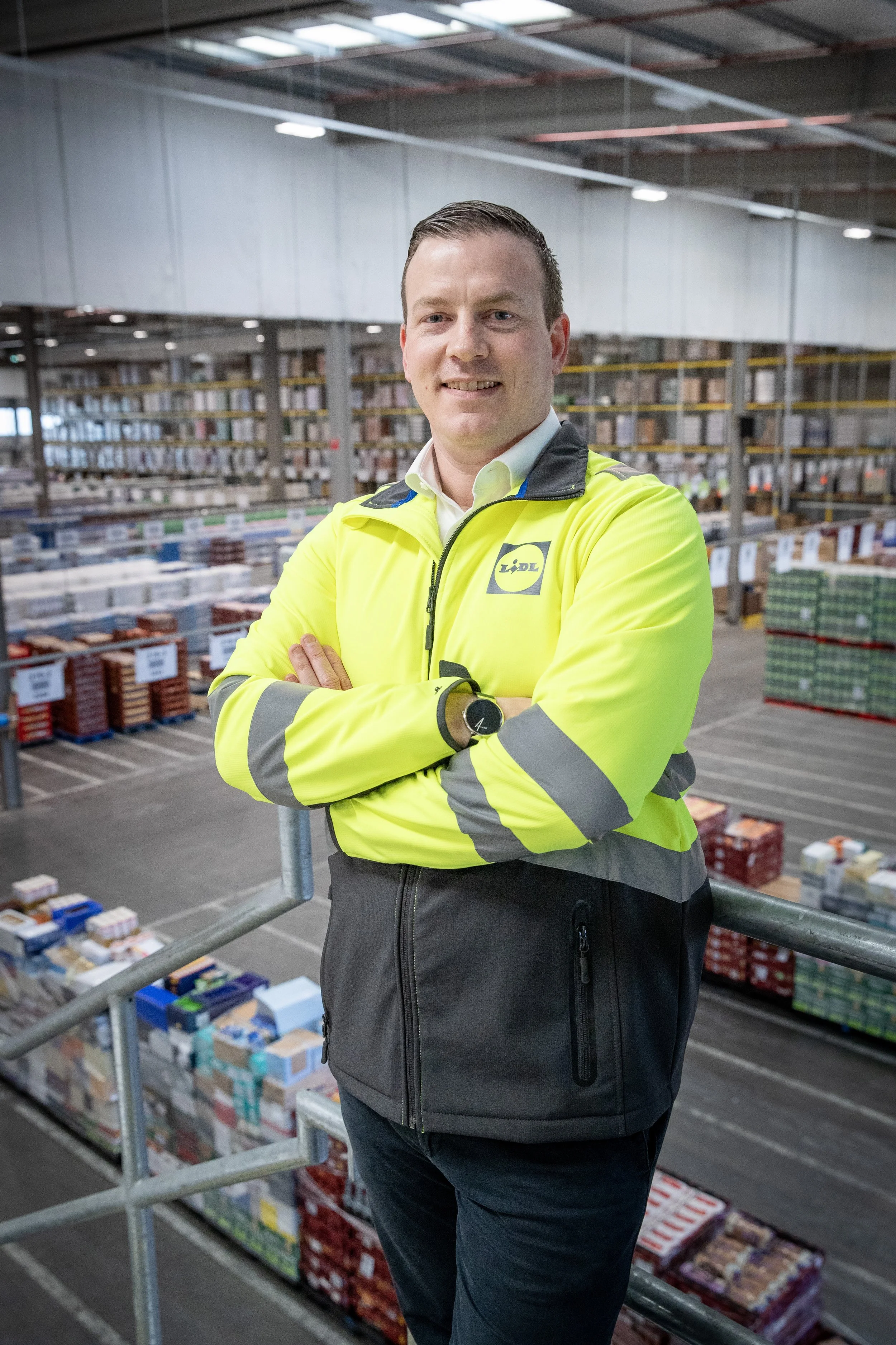 A man wearing a high-visibility yellow jacket with reflective stripes, standing in a warehouse with shelves stacked with products, arms crossed, smiling at the camera.