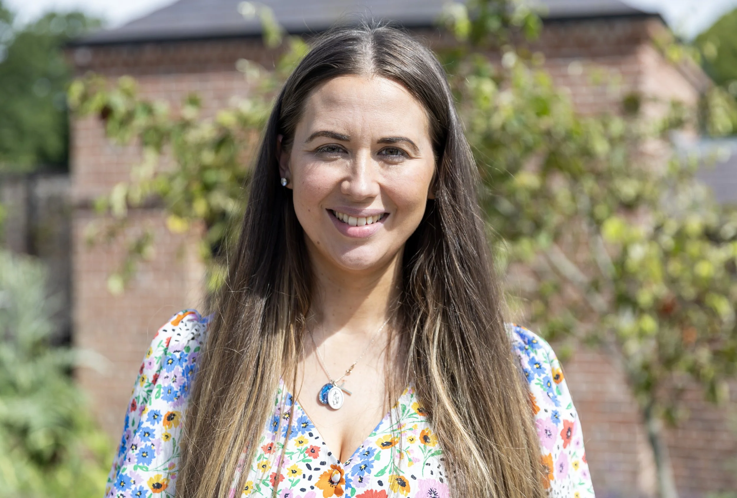A woman with long brown hair smiling outdoors in front of a brick building with trees and greenery in the background.