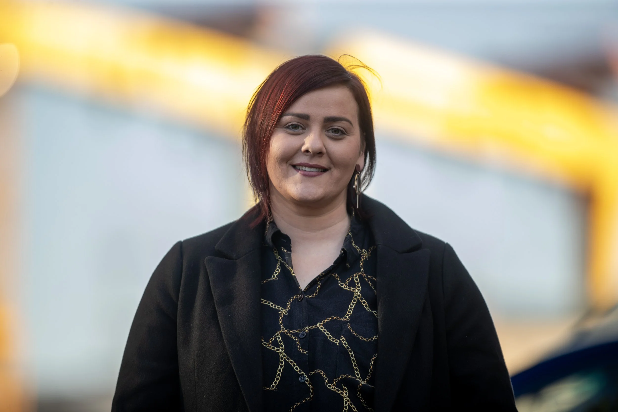 A woman with shoulder-length dark red hair, wearing a black blazer and patterned shirt, smiling outdoors with a blurry yellow structure in the background.