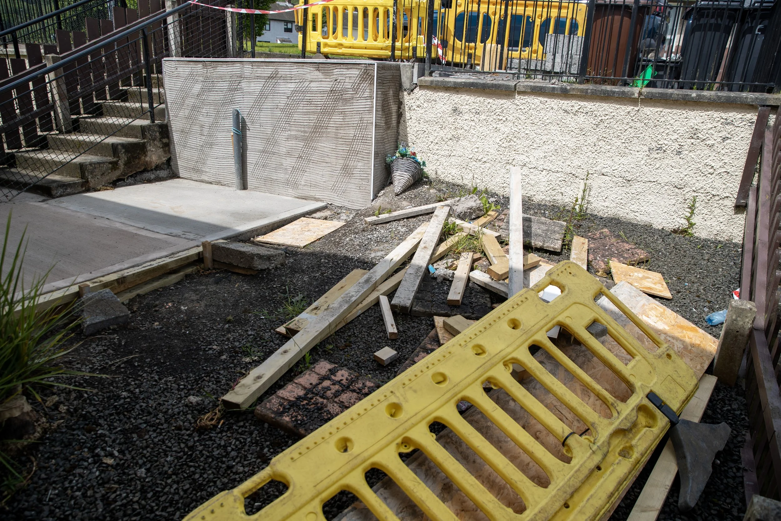 Construction site with wood planks, a yellow barrier, and concrete under renovation. There is a ramp leading to a door with safety fencing around the area.