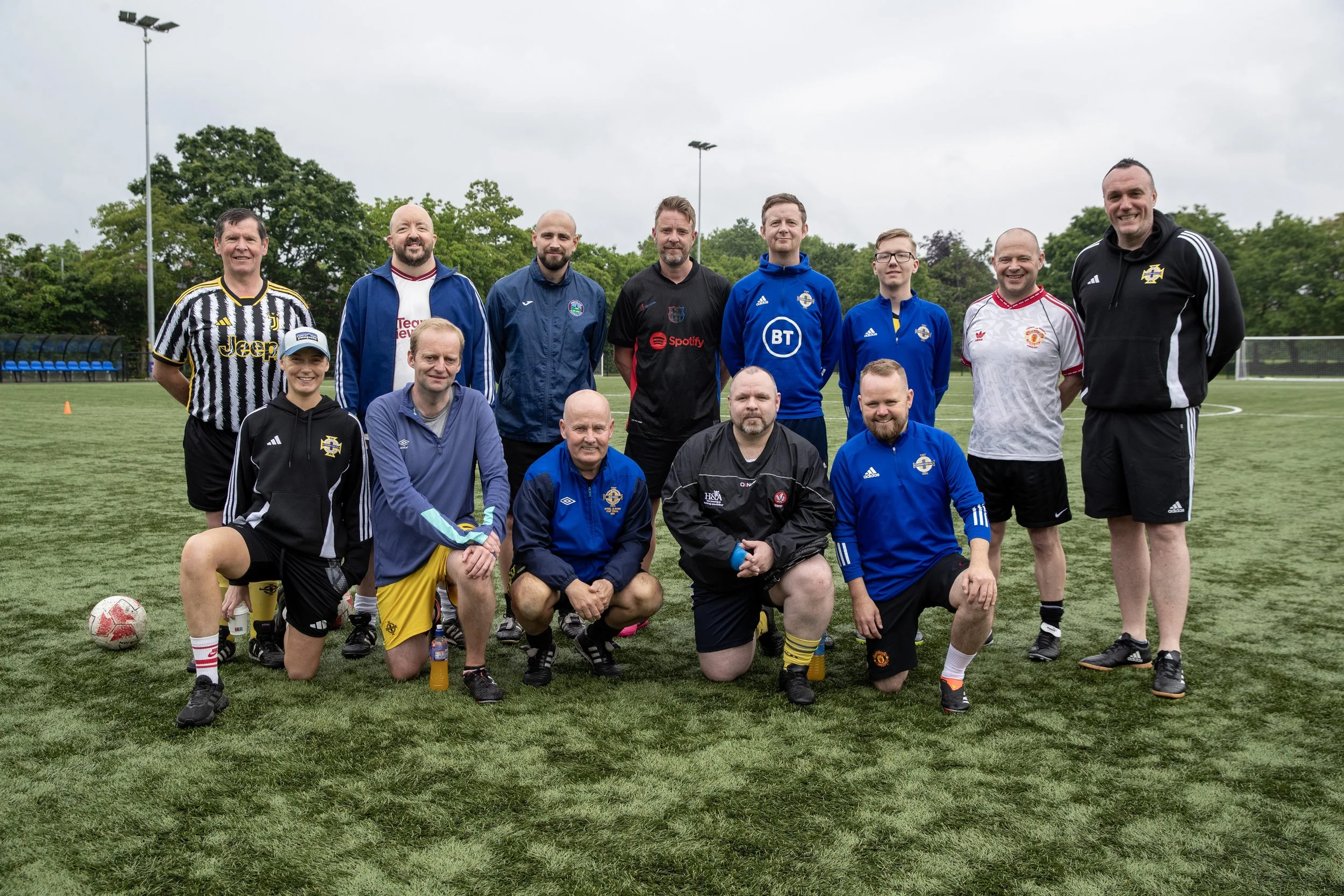 Group of 14 men and women in sports gear on a soccer field, some kneeling in front, others standing in back, on a cloudy day.