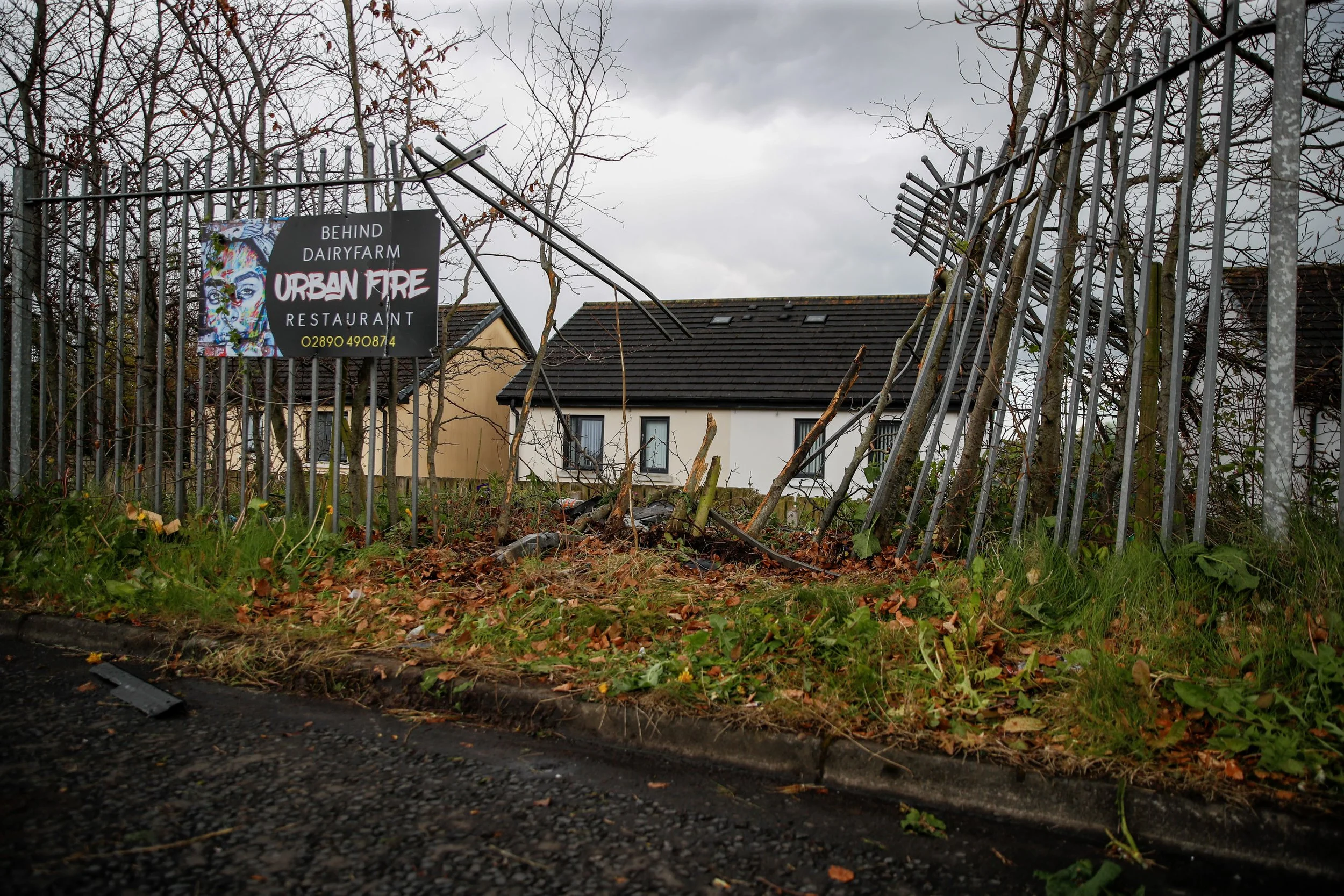 Collapsed metal fence with a sign for 'Urban Fire Restaurant' behind it, fallen tree branches on the ground, and residential houses in the background under cloudy sky.