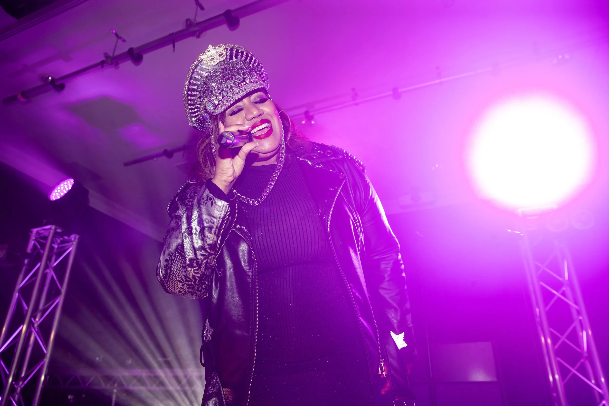 Performer wearing a decorative crown, leather jacket, and black outfit singing into a microphone on stage with purple lighting.