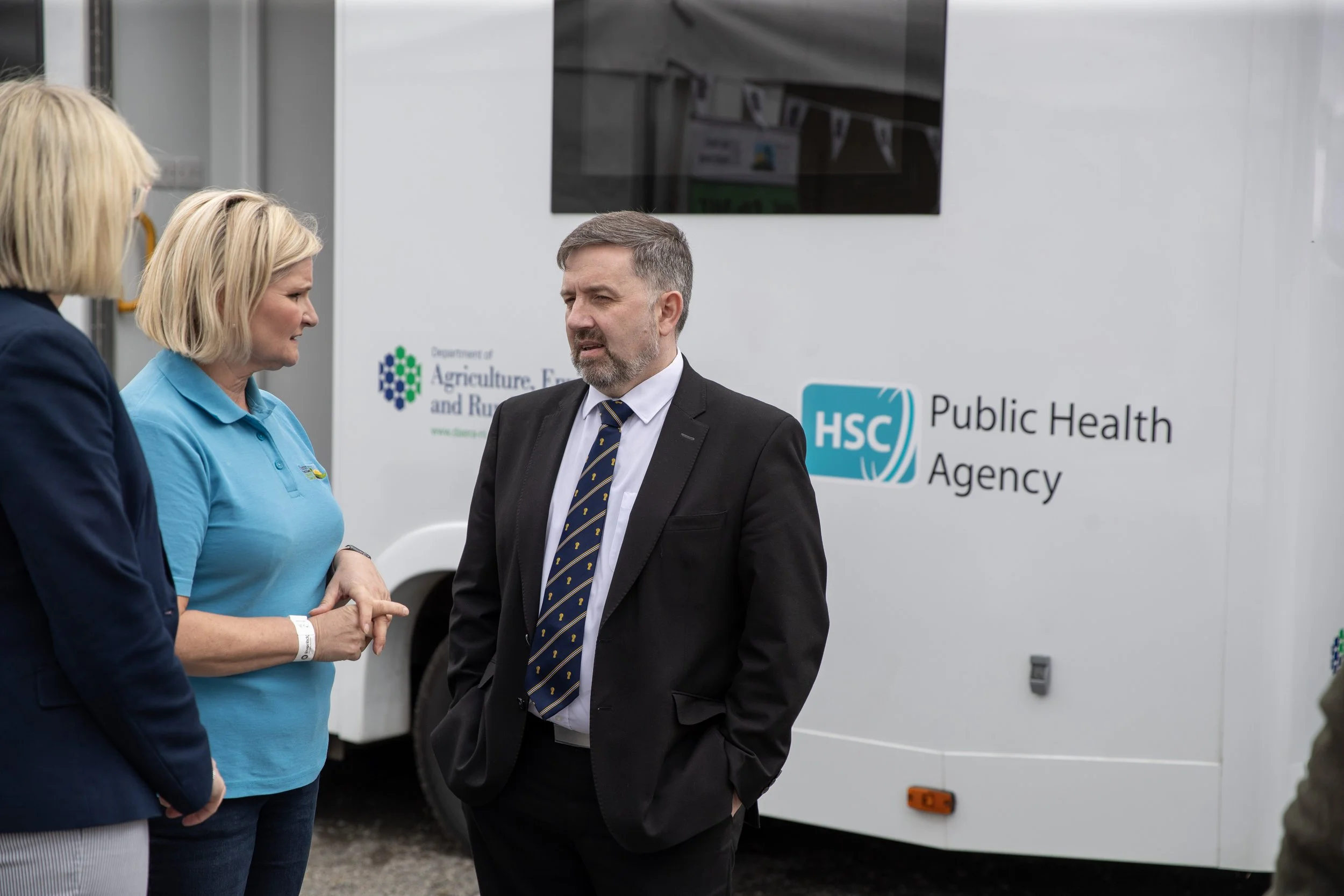 Three people having a conversation in front of a mobile health clinic with the logos of the Department of Agriculture and the Public Health Agency.