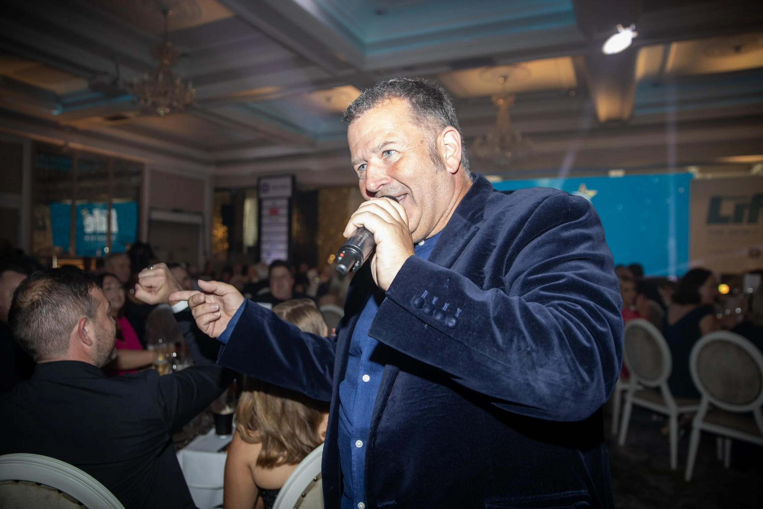 A man speaking into a microphone at a formal event with an audience seated at tables in the background.