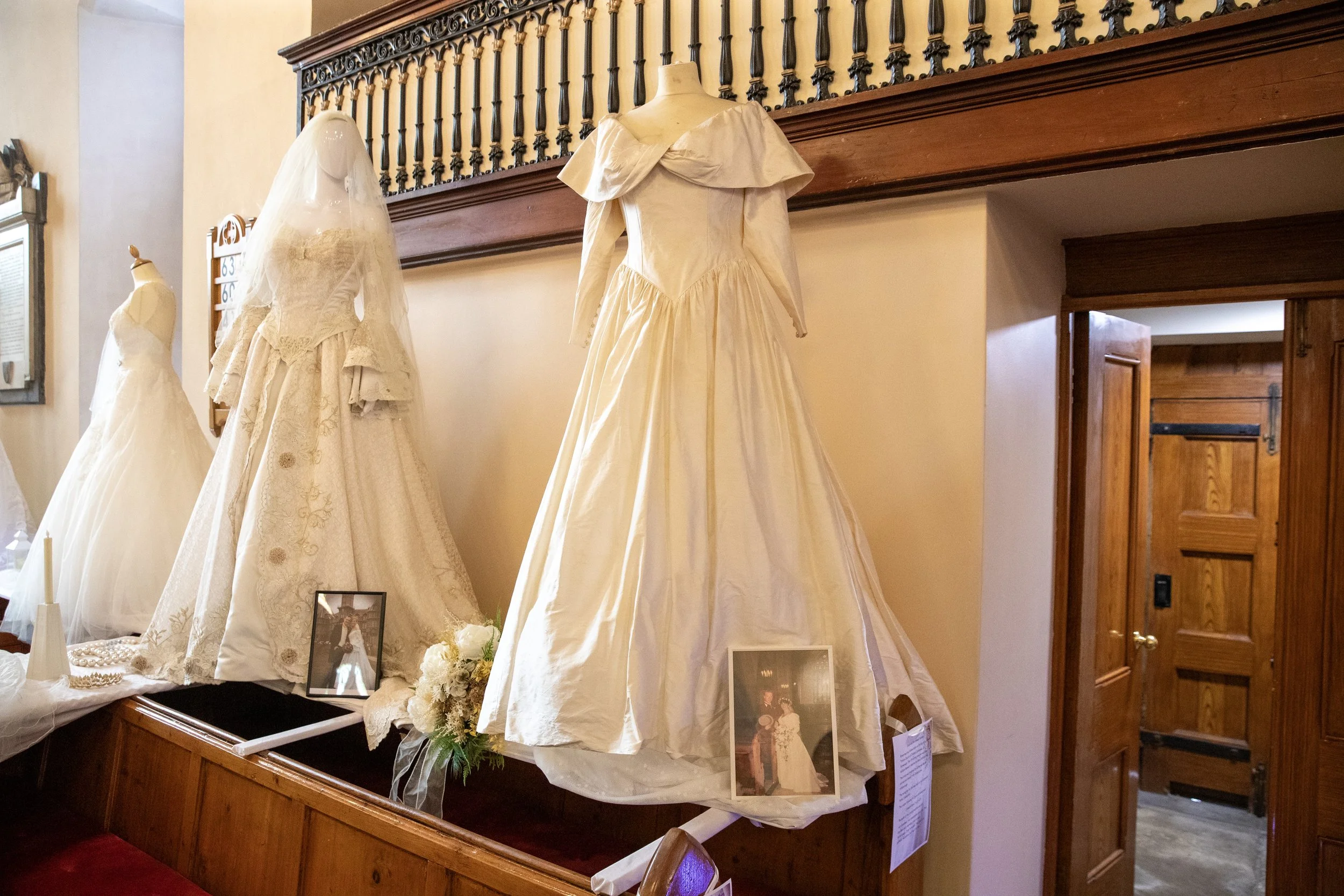 Vintage wedding dresses displayed on mannequins and a table, with framed photographs and floral arrangements, inside a room with wood accents.
