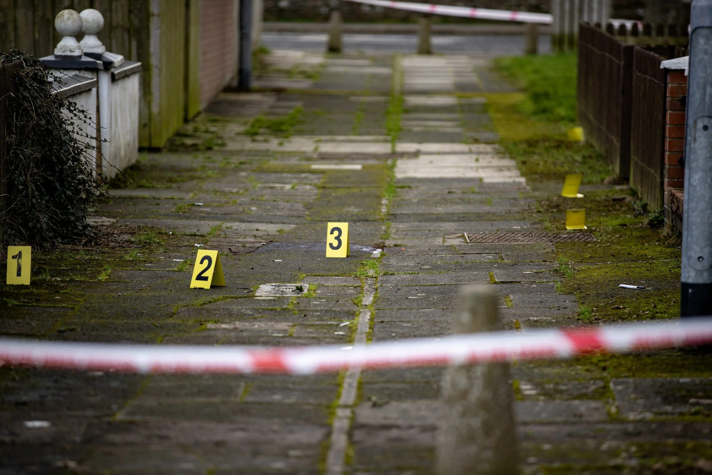 Sidewalk with yellow evidence markers numbered 1, 2, and 3, surrounded by police tape, moss and dirt on the pavement, and a residential area with fences and houses in the background.