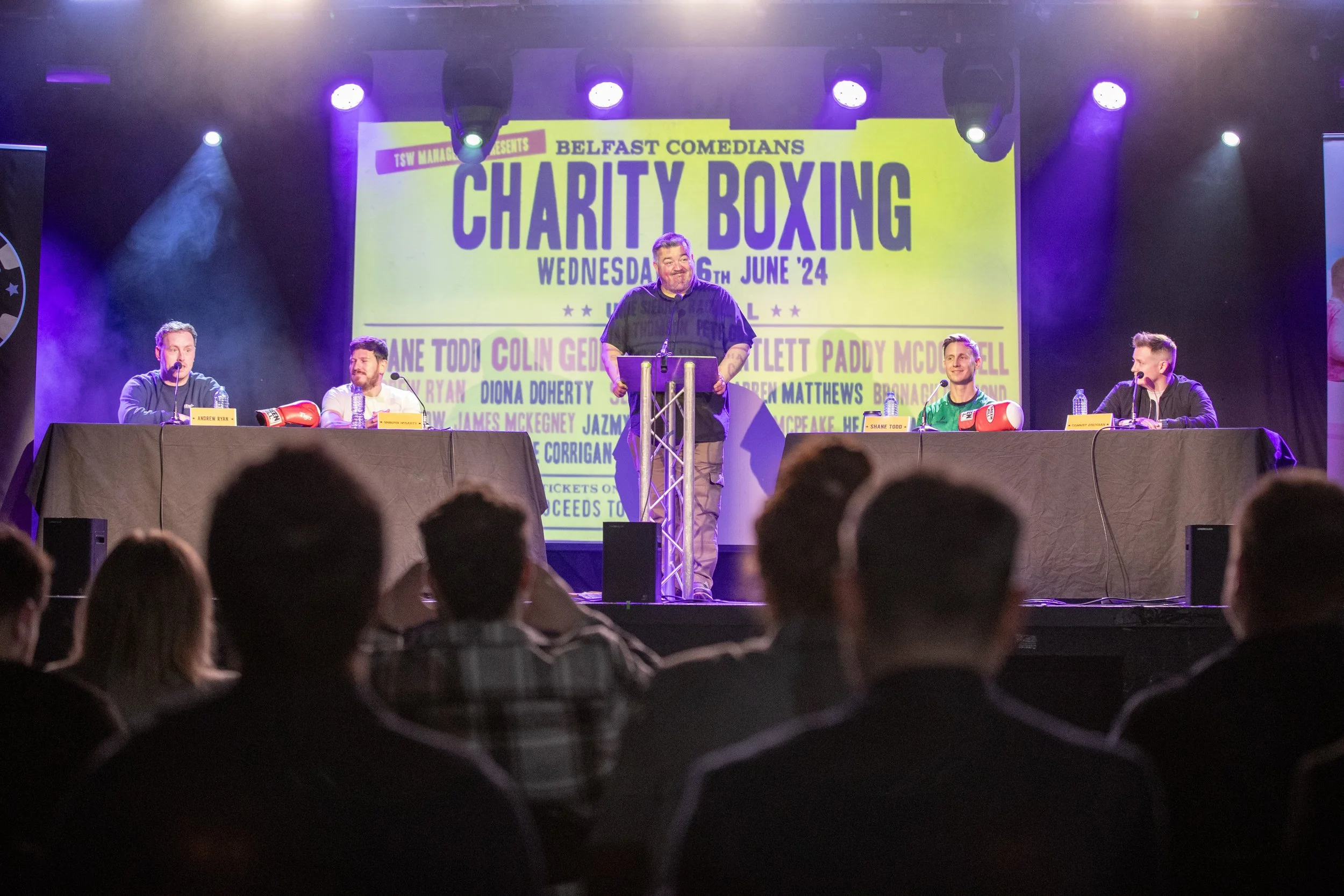 A comedy panel event about charity boxing on stage with four men, one standing at a podium, and a yellow poster behind. Audience seated in front.