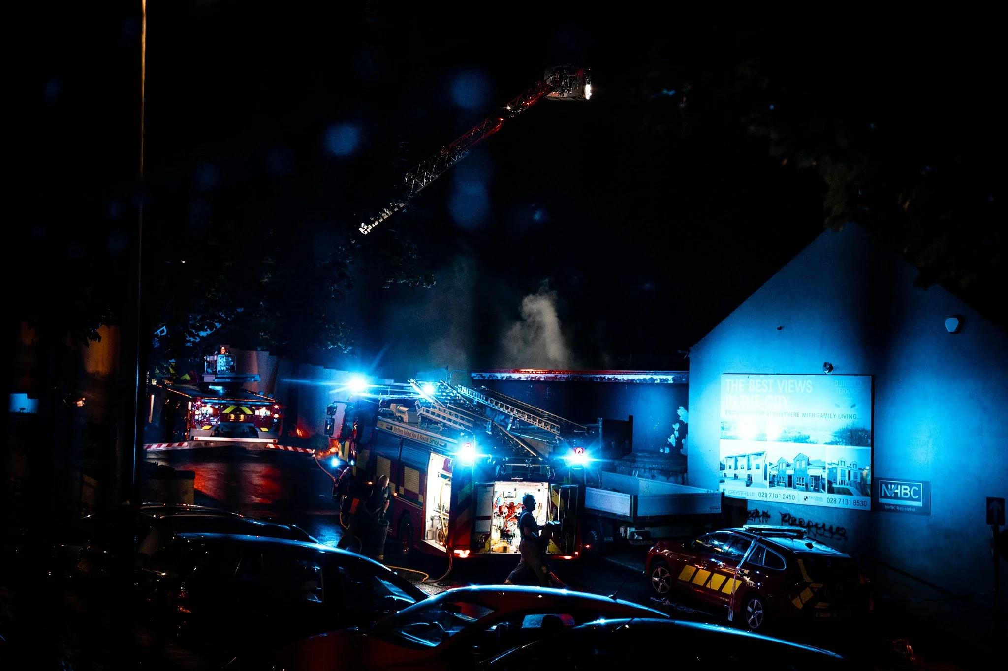 Night scene of a firefighting operation with multiple fire trucks and equipment, illuminated by blue emergency lights, on a wet street with parked cars and a billboard on a wall.