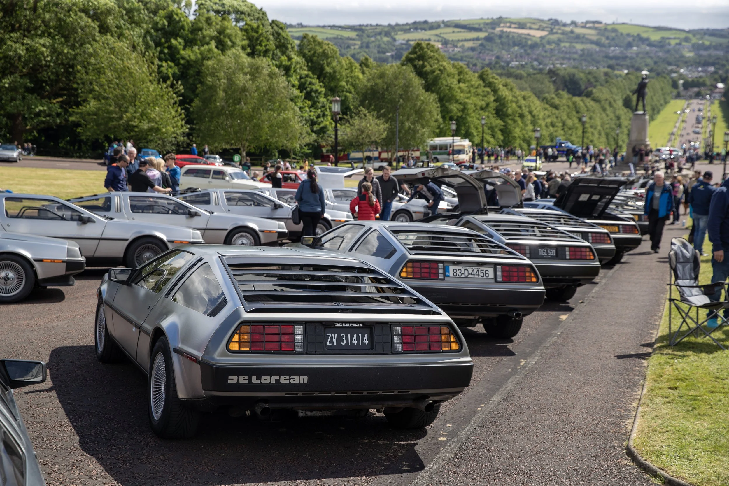 Lineup of DeLorean DMC-12 cars at a car show in an outdoor park with people walking and viewing the displays, surrounded by lush green trees and rolling hills in the background.