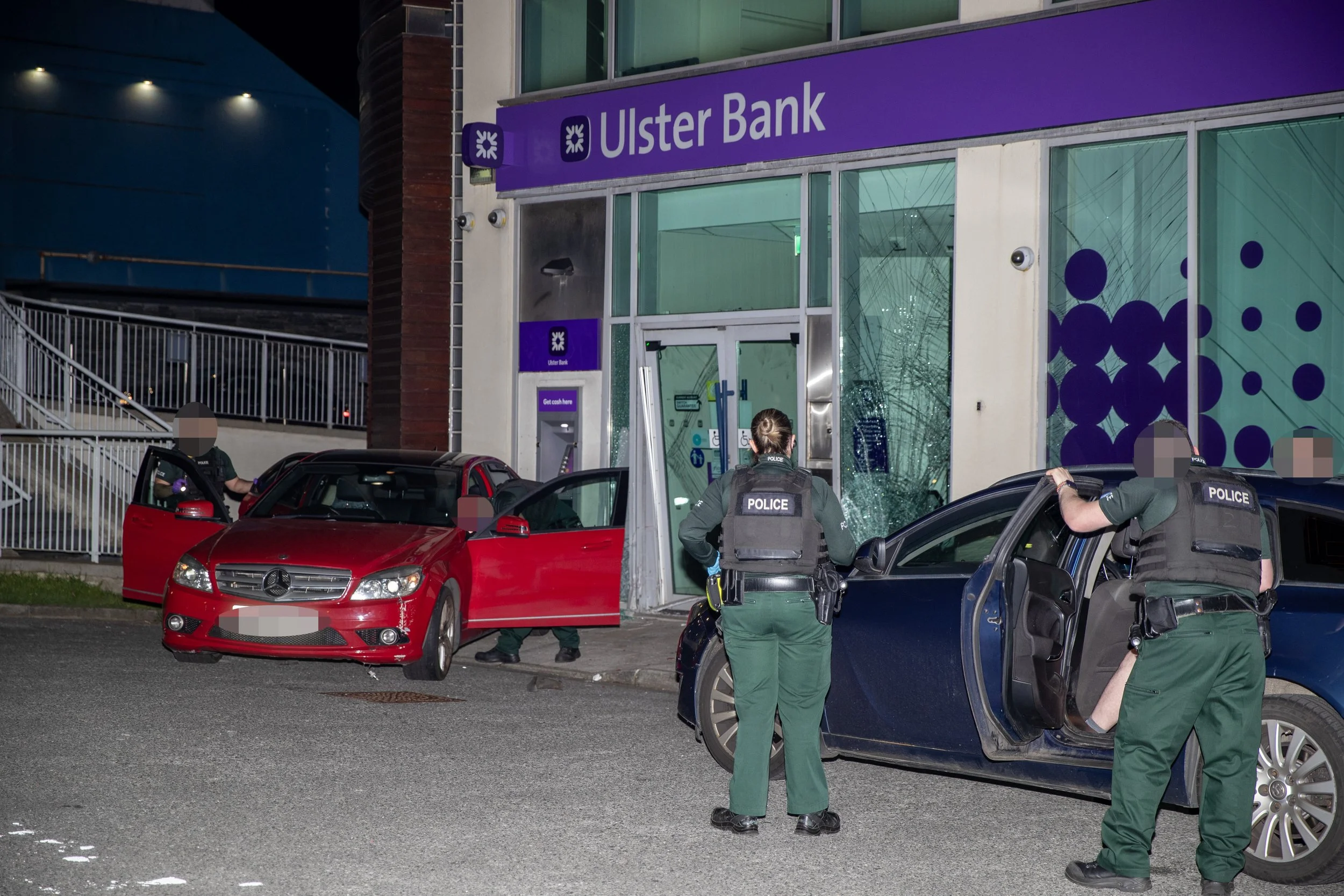 Police officers investigate after a car crashes into the storefront of Ulster Bank, which has shattered glass on the ground and a damaged door. A red Mercedes and a blue car are shown near the scene, with police officers inspecting the area during ni