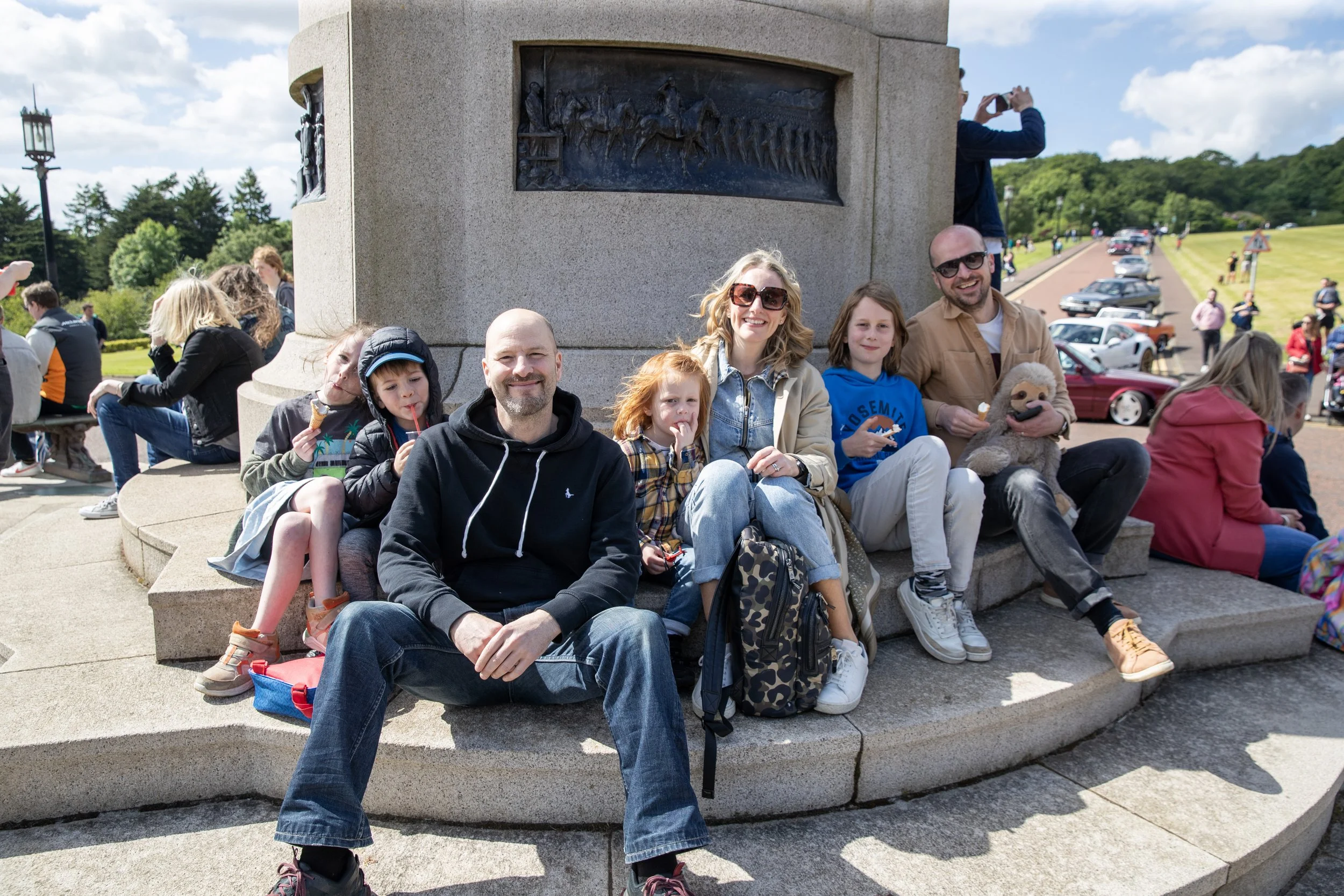 Family group sitting on a monument base outdoors on a sunny day, with cars and other people in the background.