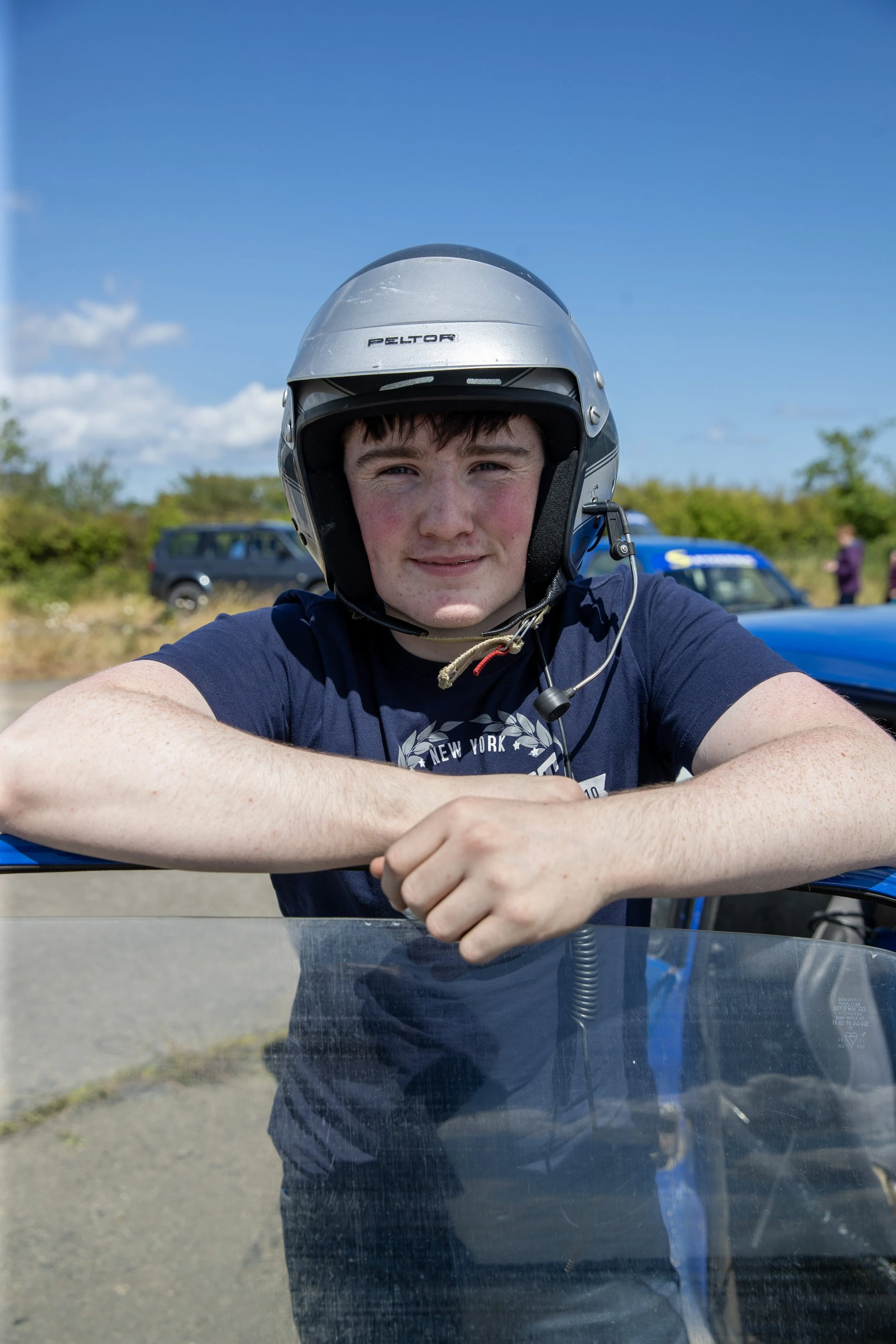 Young man wearing a motorcycle helmet and blue T-shirt, resting his arms on a motorcycle's windshield outdoors on a sunny day with cars and trees in the background.