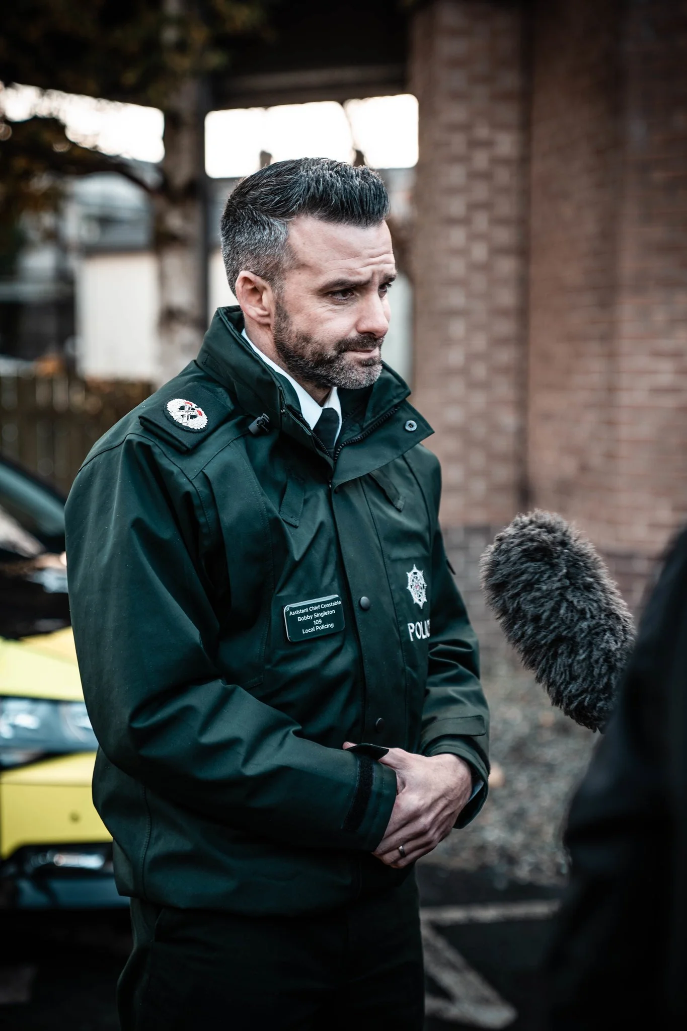 A police officer being interviewed outdoors, standing in front of a brick building, with a blurred police car and trees in the background.